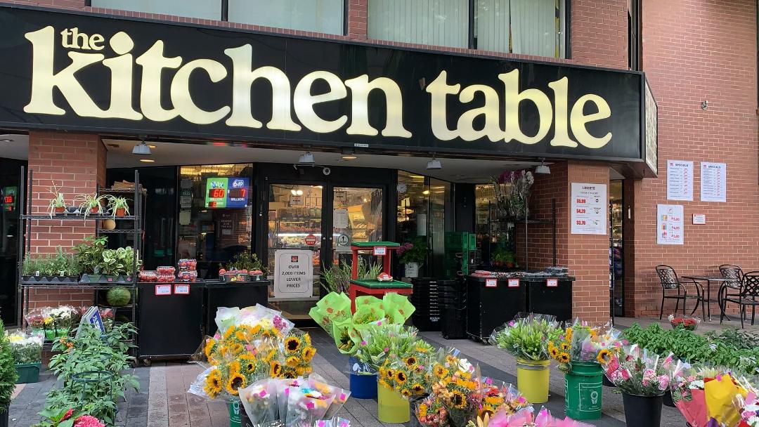 Colourful flowers and flowerpots outside The Kitchen Table grocery store with huge lettering.