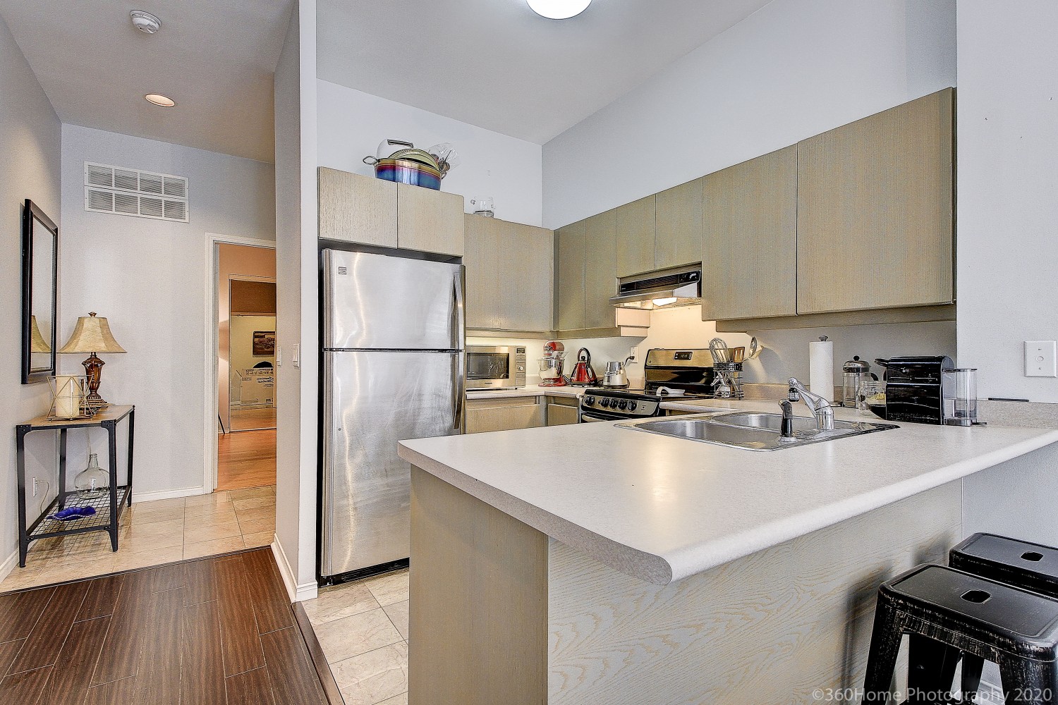 Breakfast bar with white counter-top, wood base and 2 black stools with kitchen in background.