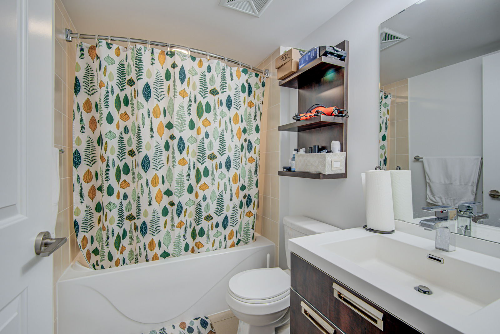 Bathroom with floral shower curtain, white tub and sink, large mirror and shelves with hairdryer and toiletries.