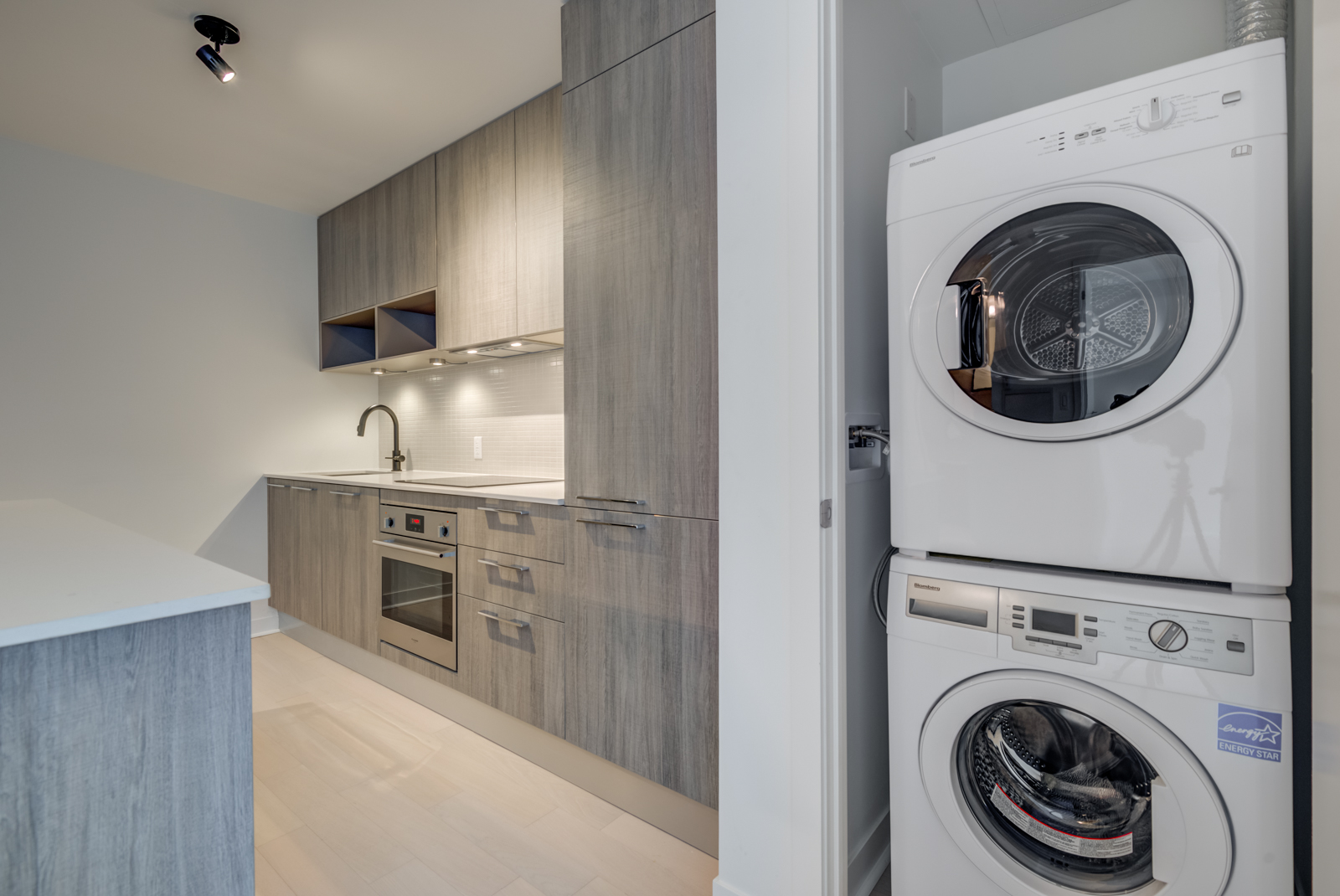 Open laundry closet showing stacked white washer and dryer plus kitchen to its left.