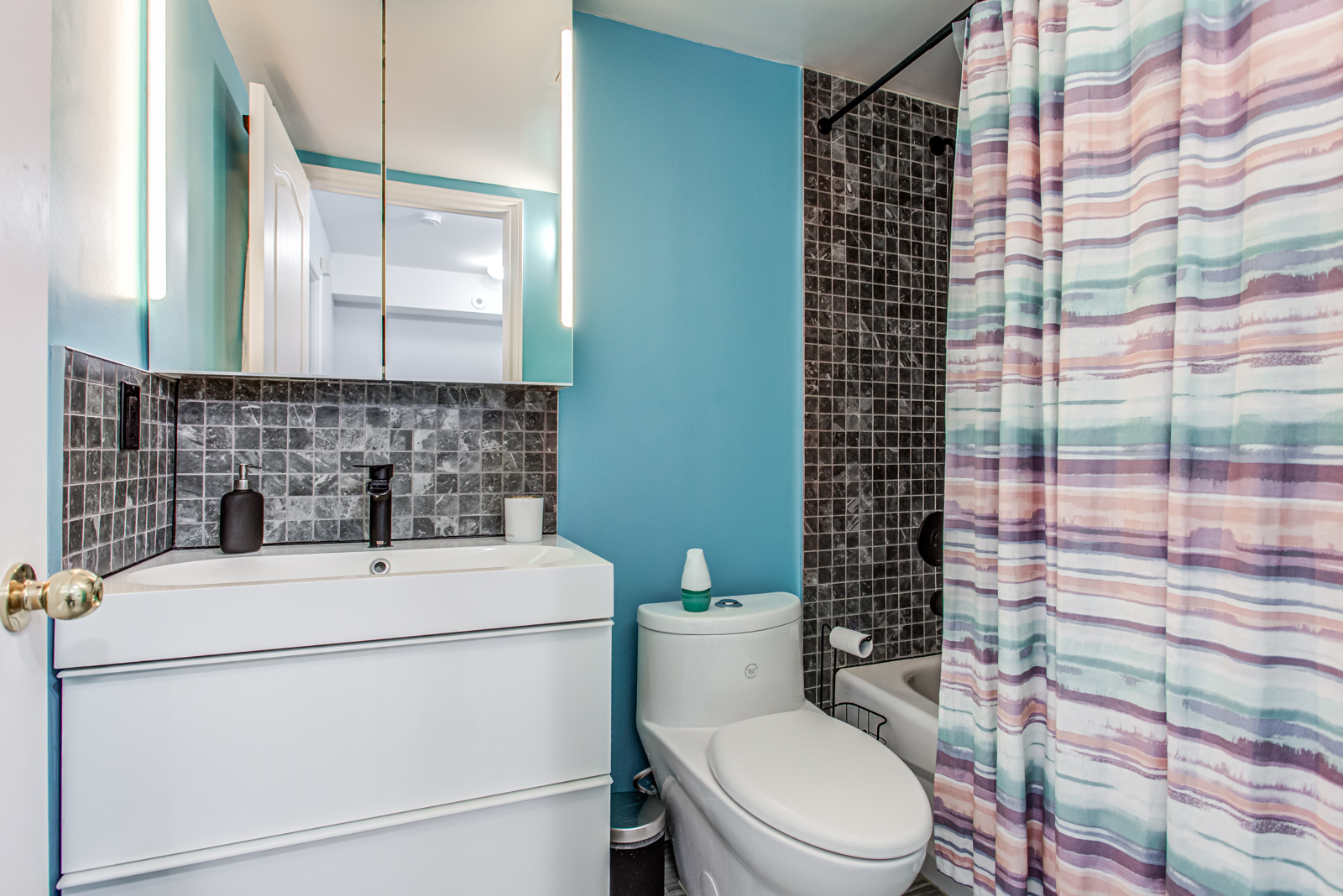 Newly-renovated bathroom with bright blue walls, small black tiles, colourful curtains and black faucet.