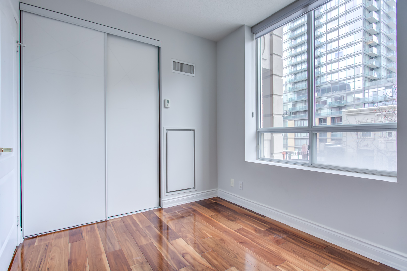 Small empty bedroom with shiny brown hardwood floors and large window showing building.