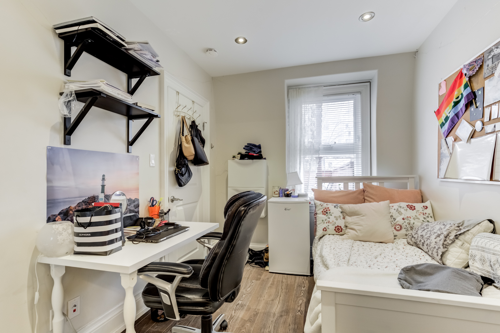 Bedroom with bed, messy desk, black wall-mounted shelves and white mini-fridge.