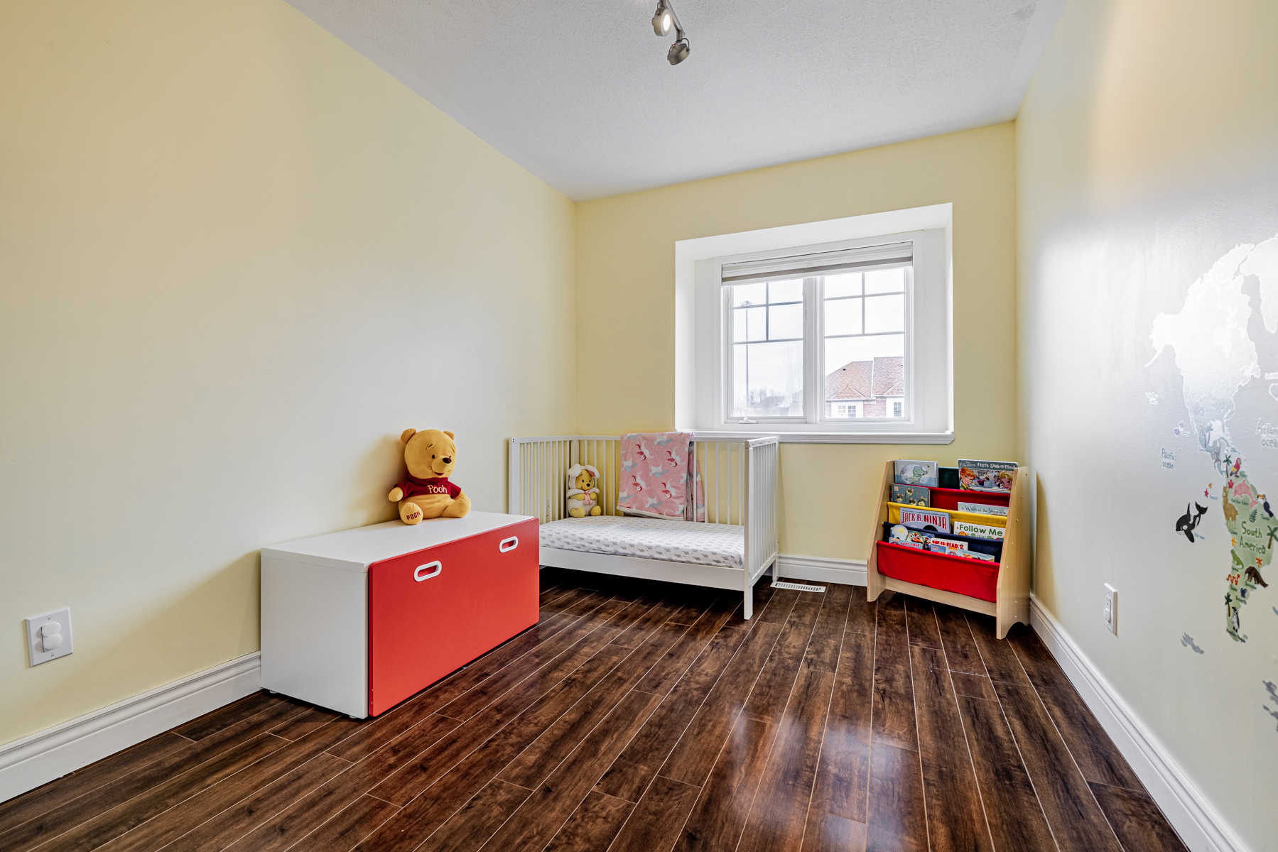 Bedroom nursery with crib, Winnie the Poo stuffy, bookshelf, yellow walls and laminate floors.