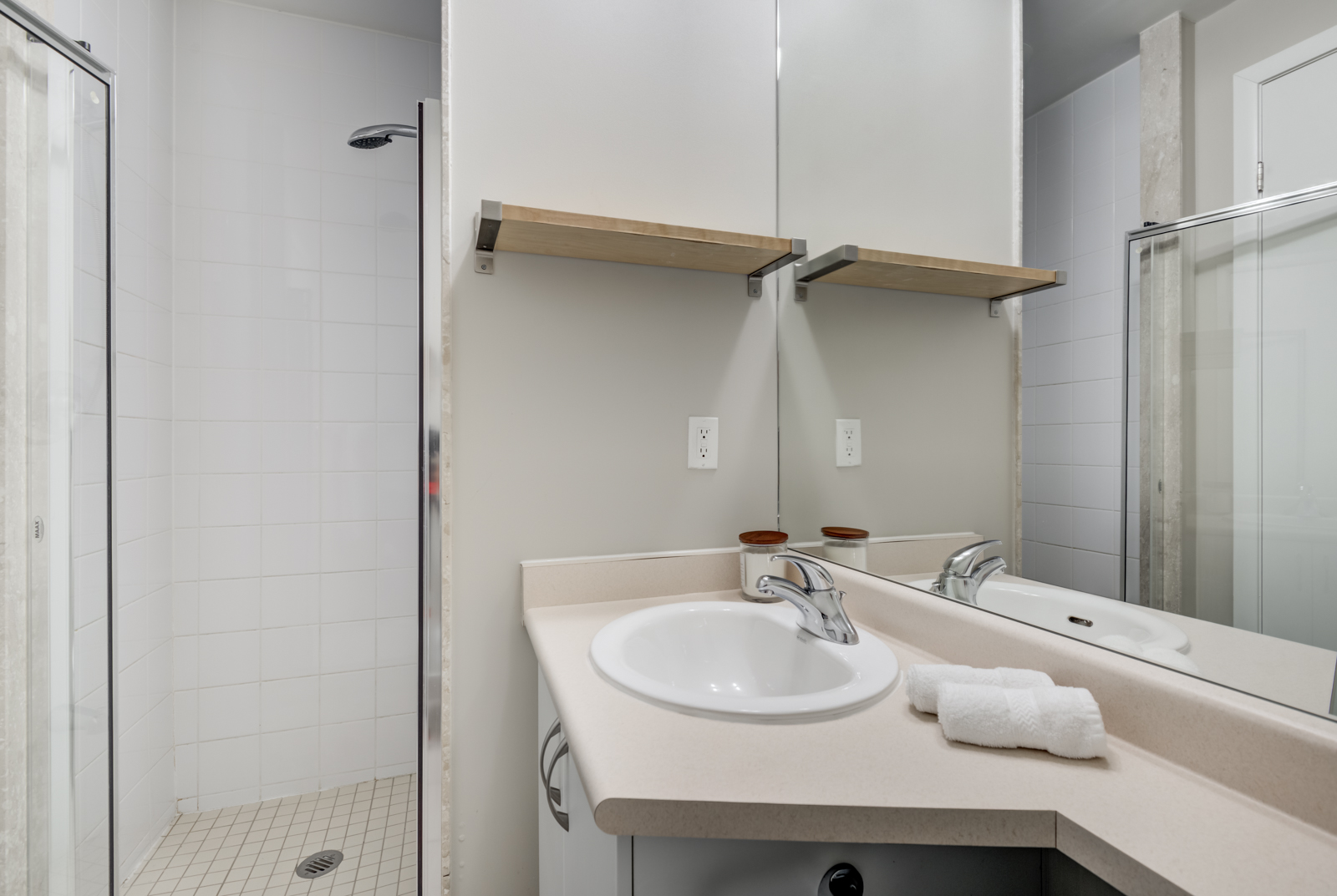 Small bathroom with oval sink, silver faucet, wooden shelf, walk-in shower and large mirror.