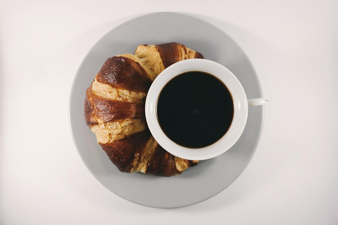 Cup of coffee with croissant wrapped around cup on simple white background.
