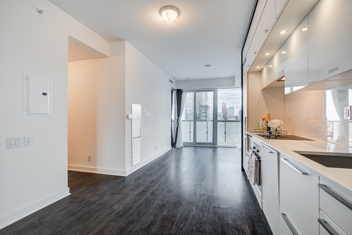 Empty condo with dark laminate floors, white kitchen and large windows.