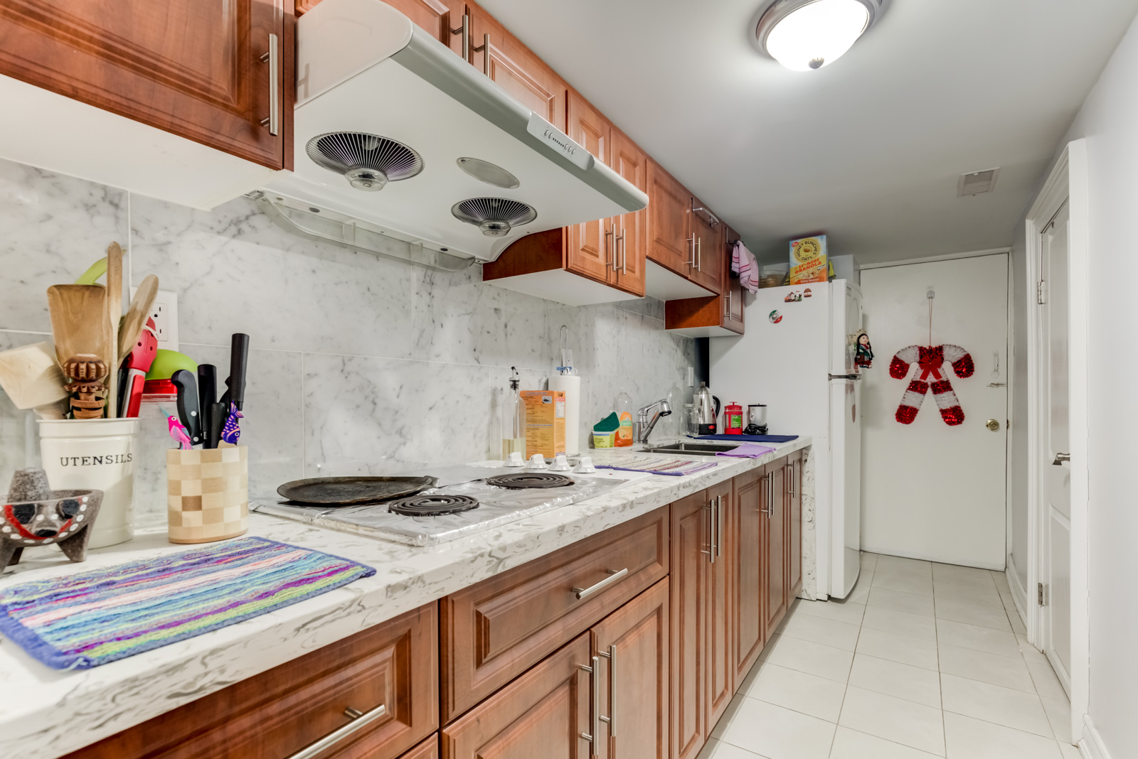 Linear kitchen with brown wooden cabinets, large-tiled backsplash and gray counters.