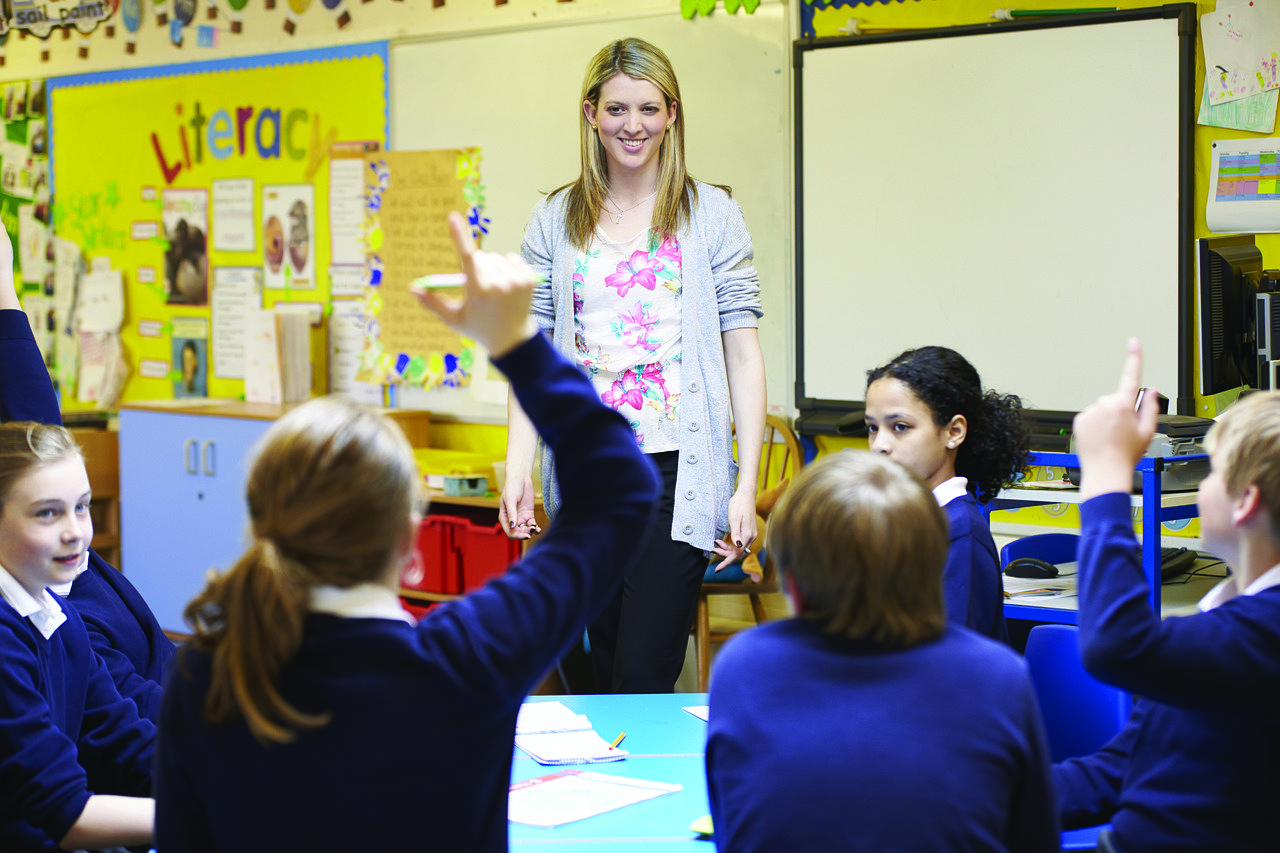 Elementary school kids in blue uniform raising hand for female teacher.