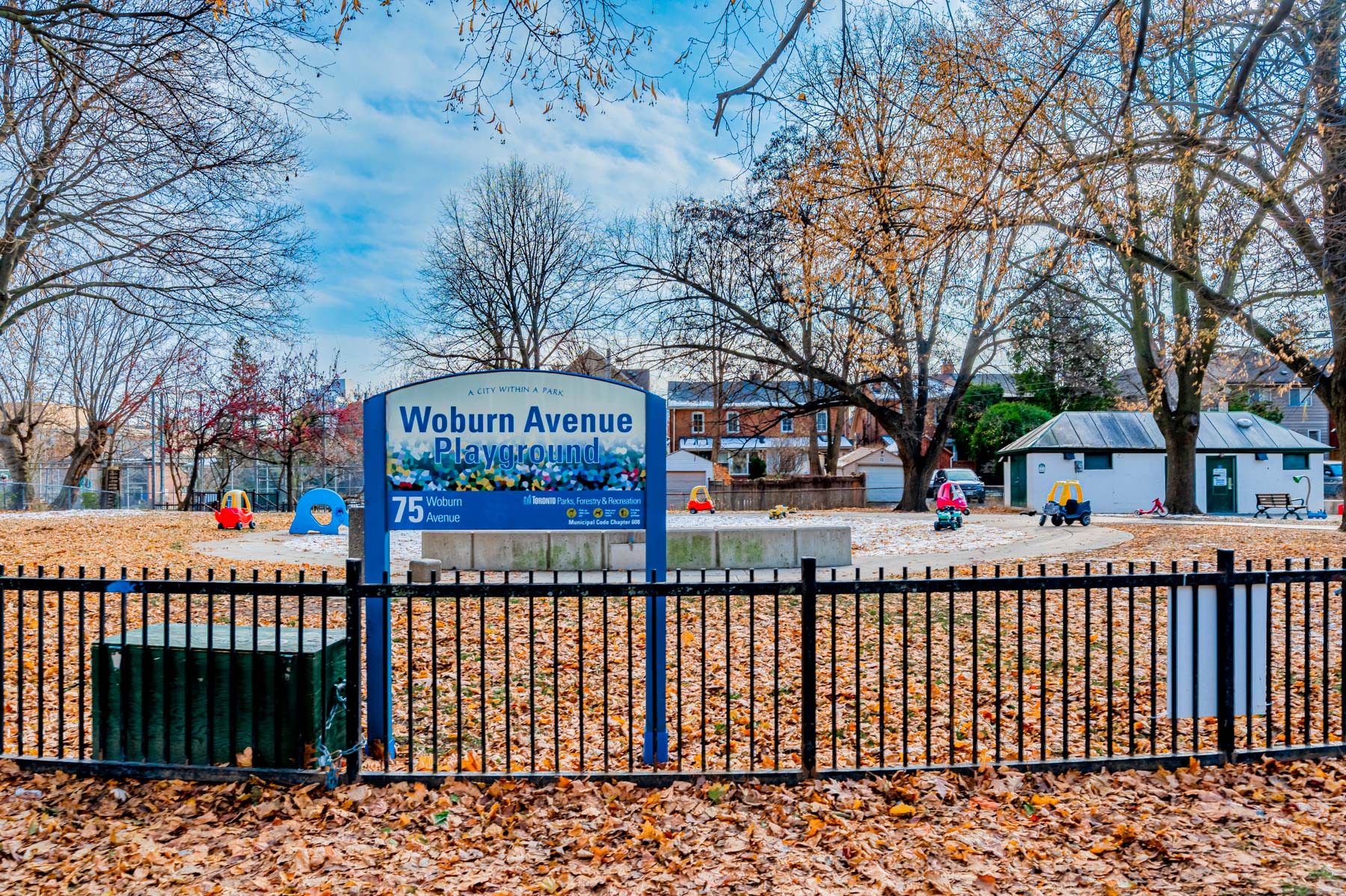 Iron fence, sign, trees and playground – Woburn Ave Playground.