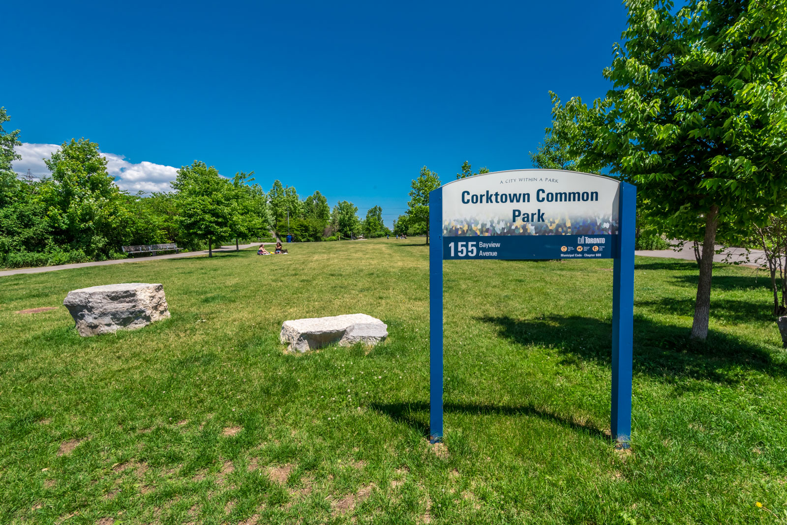 Picture showing Corktown Common Park and people. We see the park sign and also beautiful green grass.
