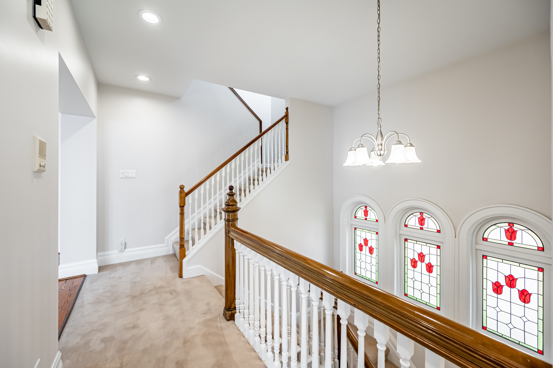 House stairway with shiny wood banisters, silver chandelier, pot-lights and stained glass windows.