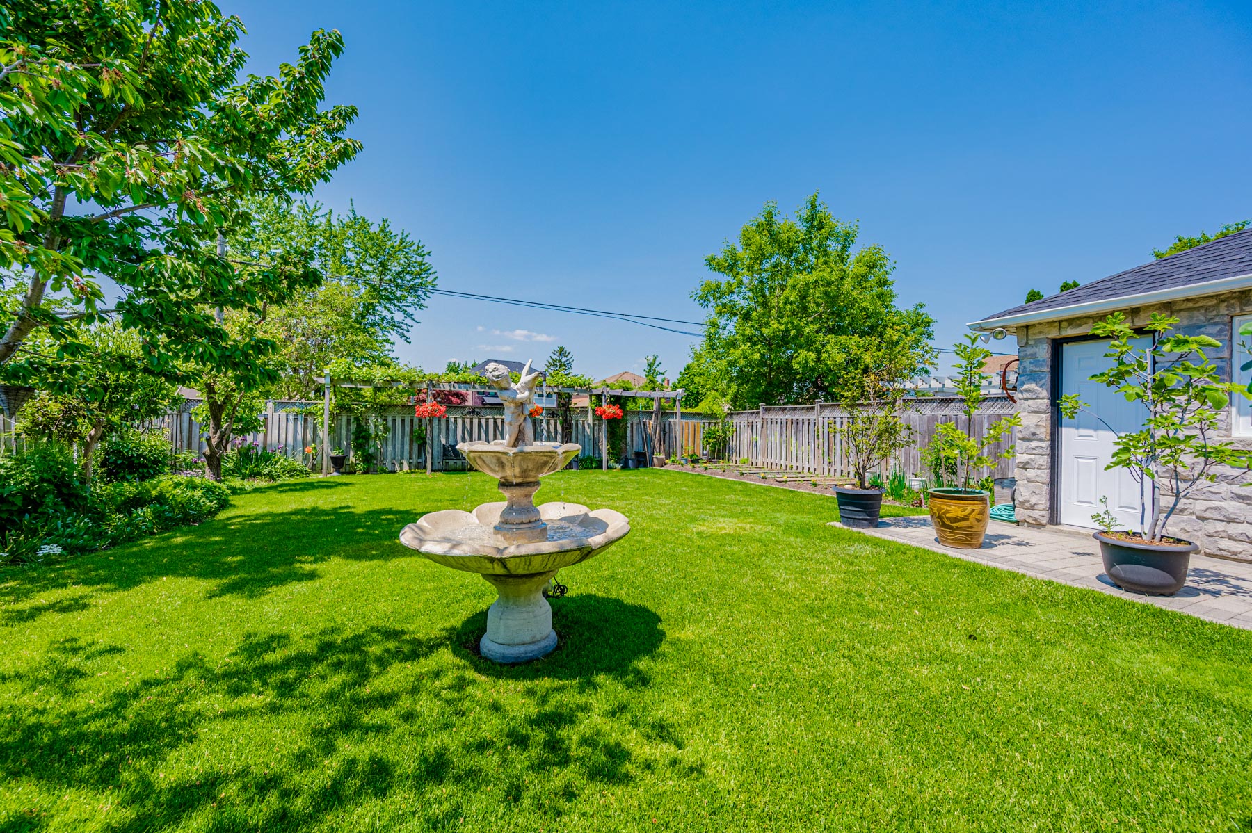Backyard with fountain, trees, shrubs, garden plots and high fence.