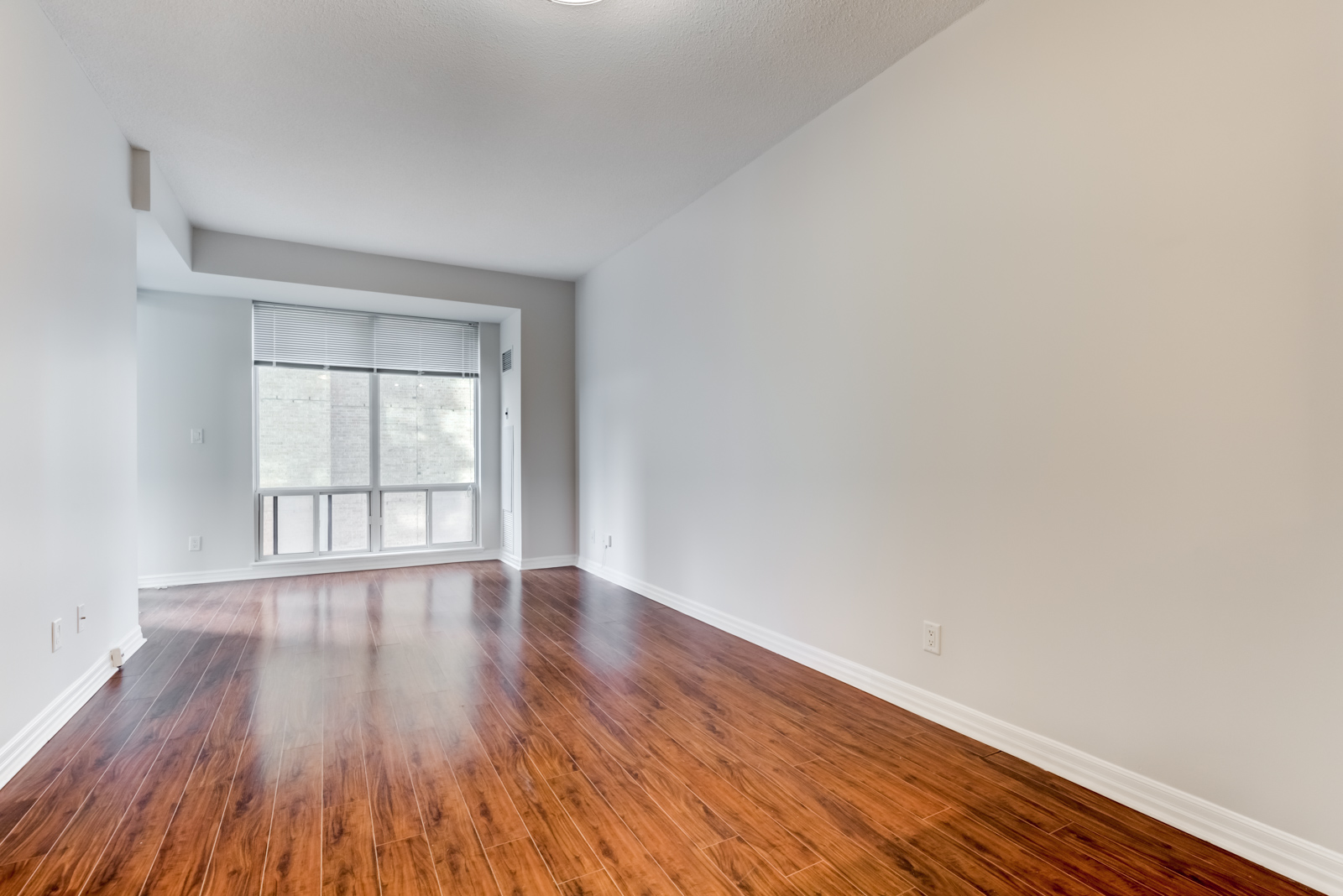 Empty living and dining room of 300 Bloor St E Unit 1809 with dark brown laminate floors shining with light from window.