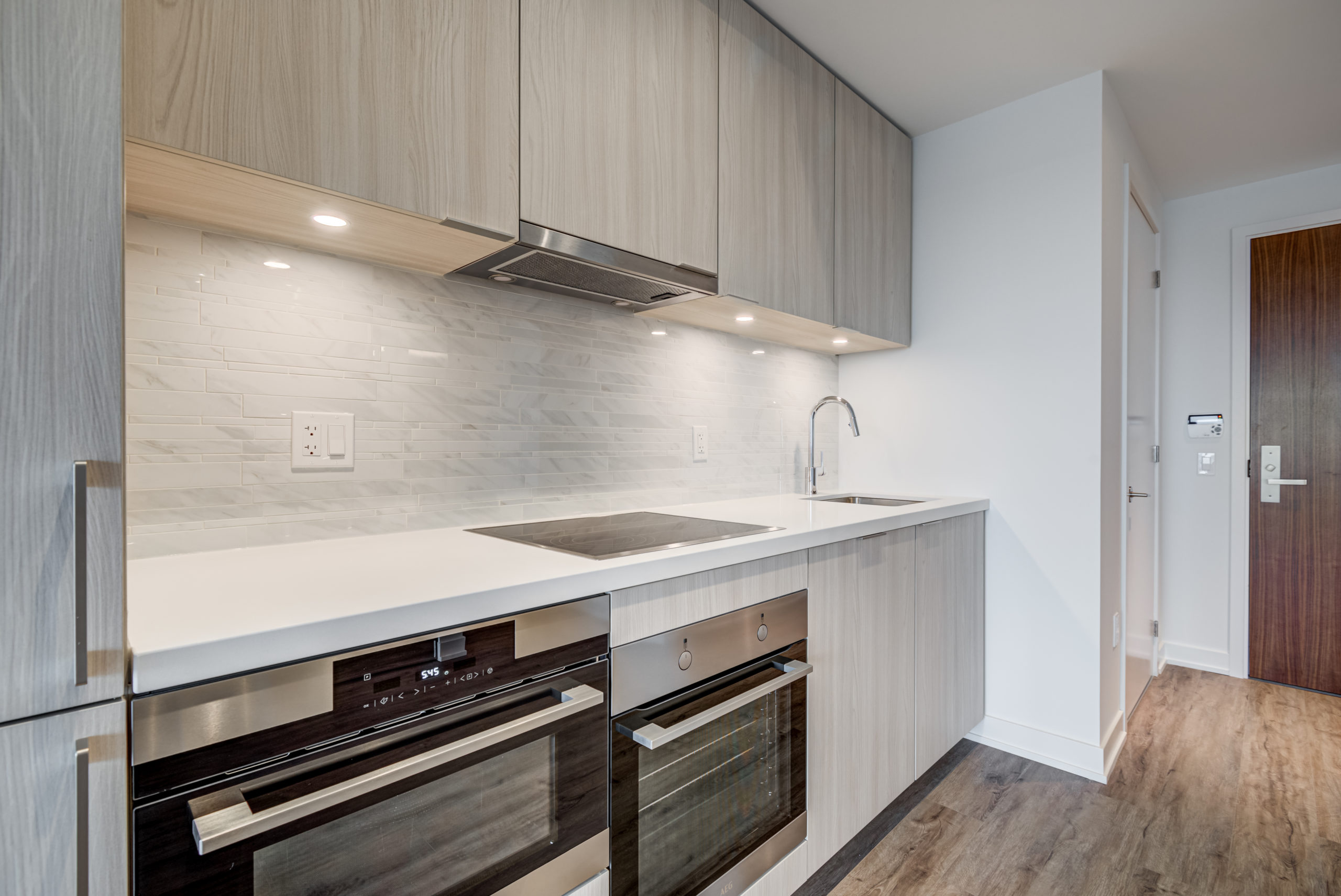 Condo kitchen with white counters, large cabinets with lights and glazed backsplash.