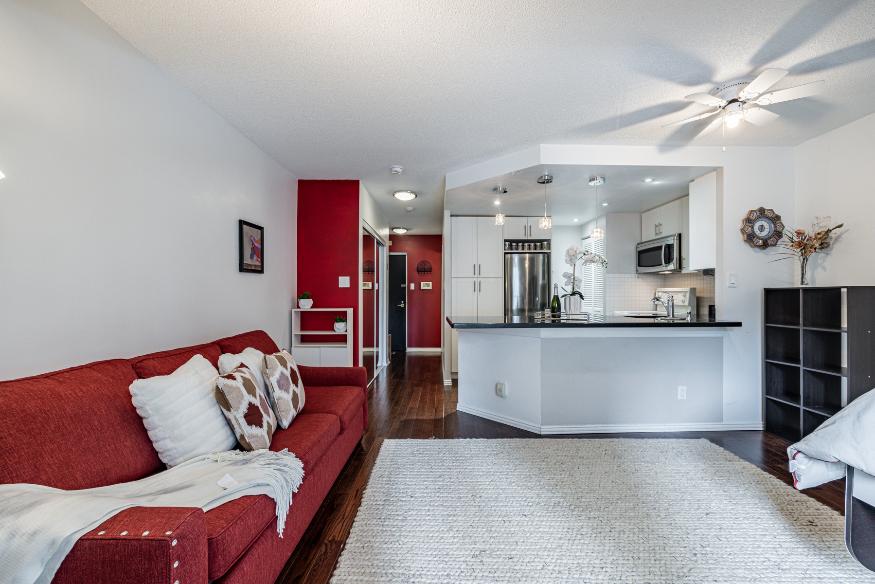 Condo living room containing ceiling fan with light bulb and kitchen with pendant lights and pot-lights.