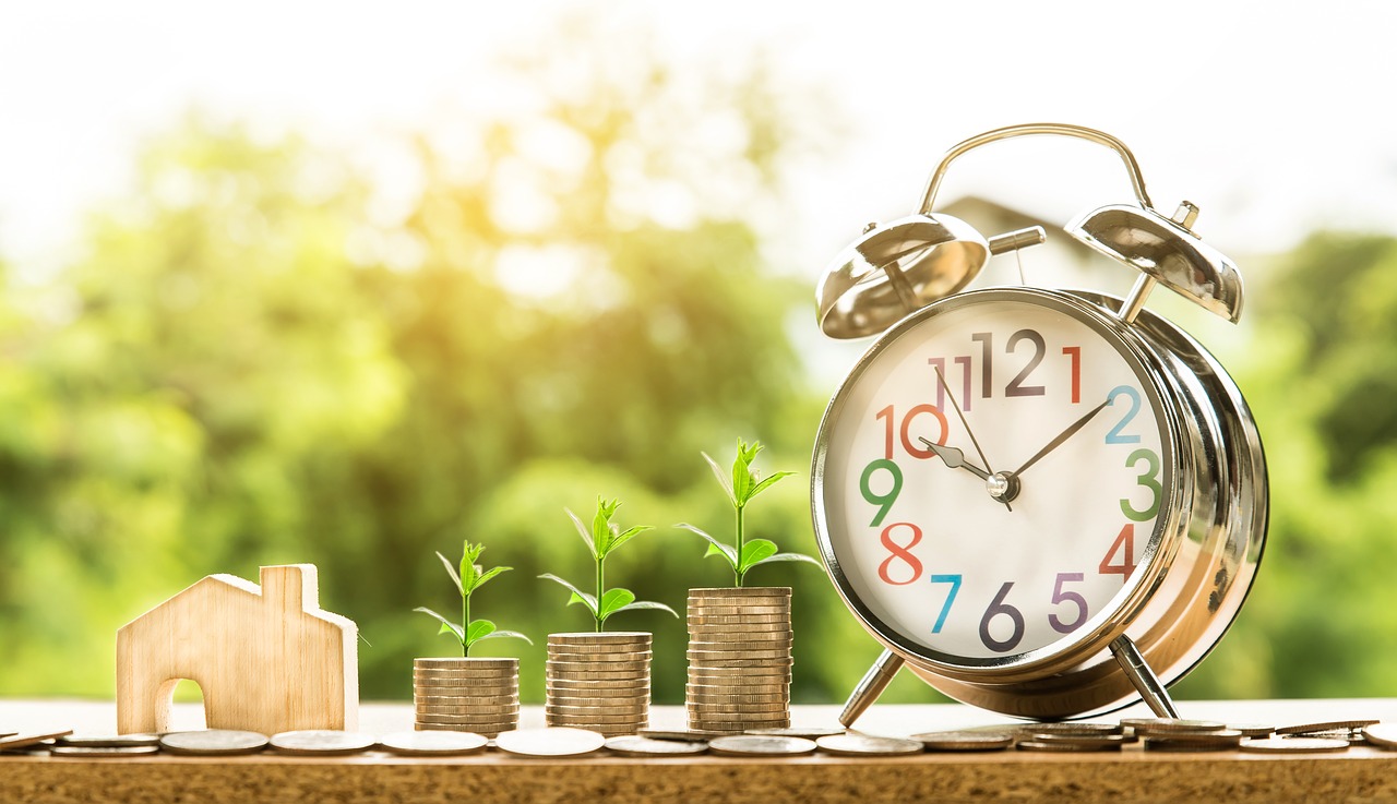 Analog clock, stacks of coin and model wooden house.