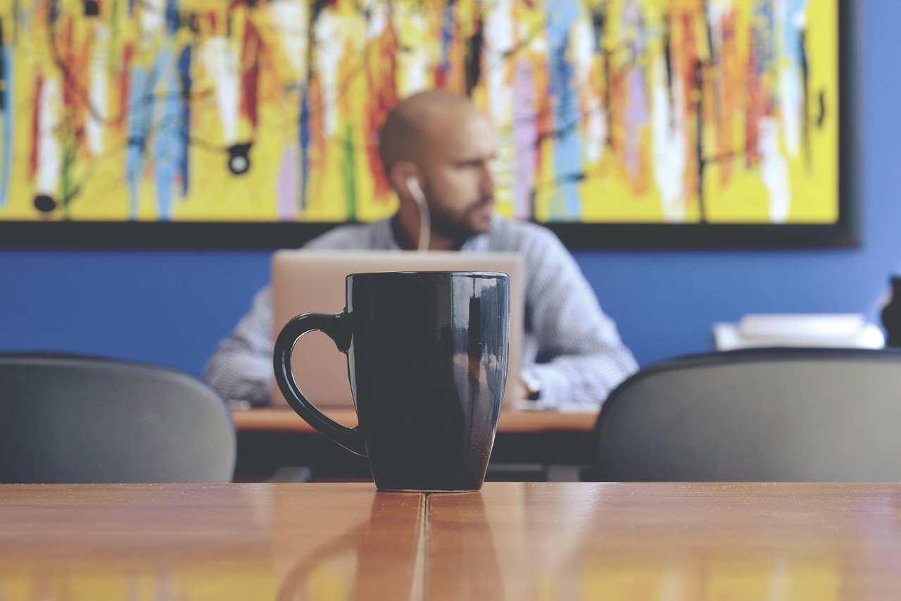 Black coffee mug in foreground and man with laptop in background.