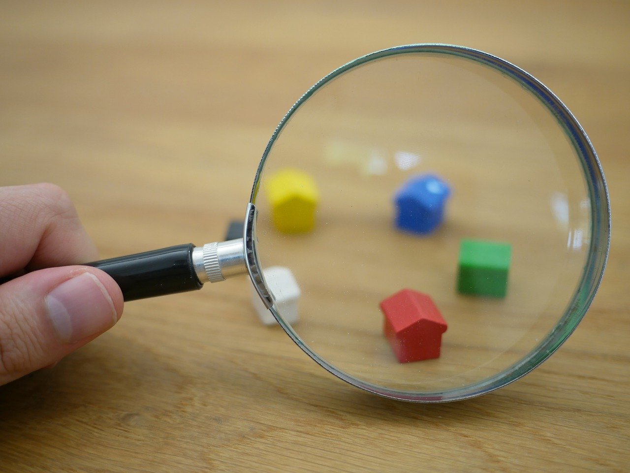 Hand holding magnifying glass in front of Monopoly house pieces.
