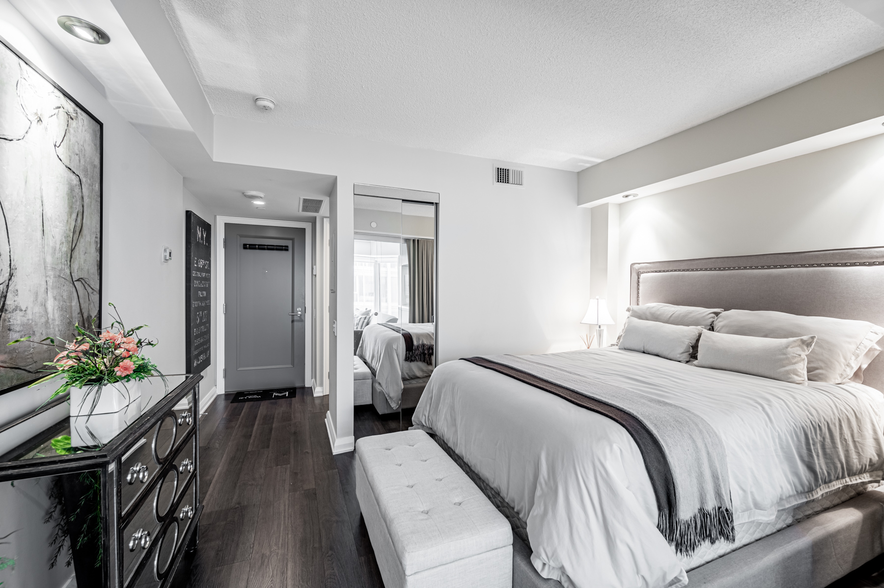 Condo bedroom with storage bench, painting and dresser with colourful flowers.