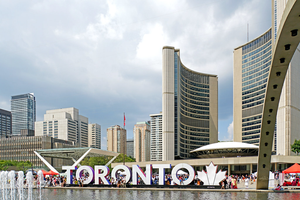Toronto sign in foreground and City Hall and luxury real estate condos in background.