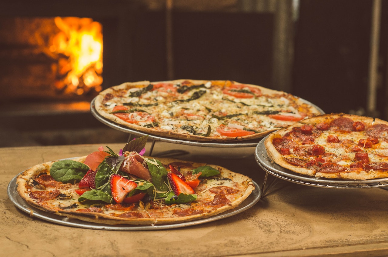 Three pizzas with several ingredients and pizza oven in background.