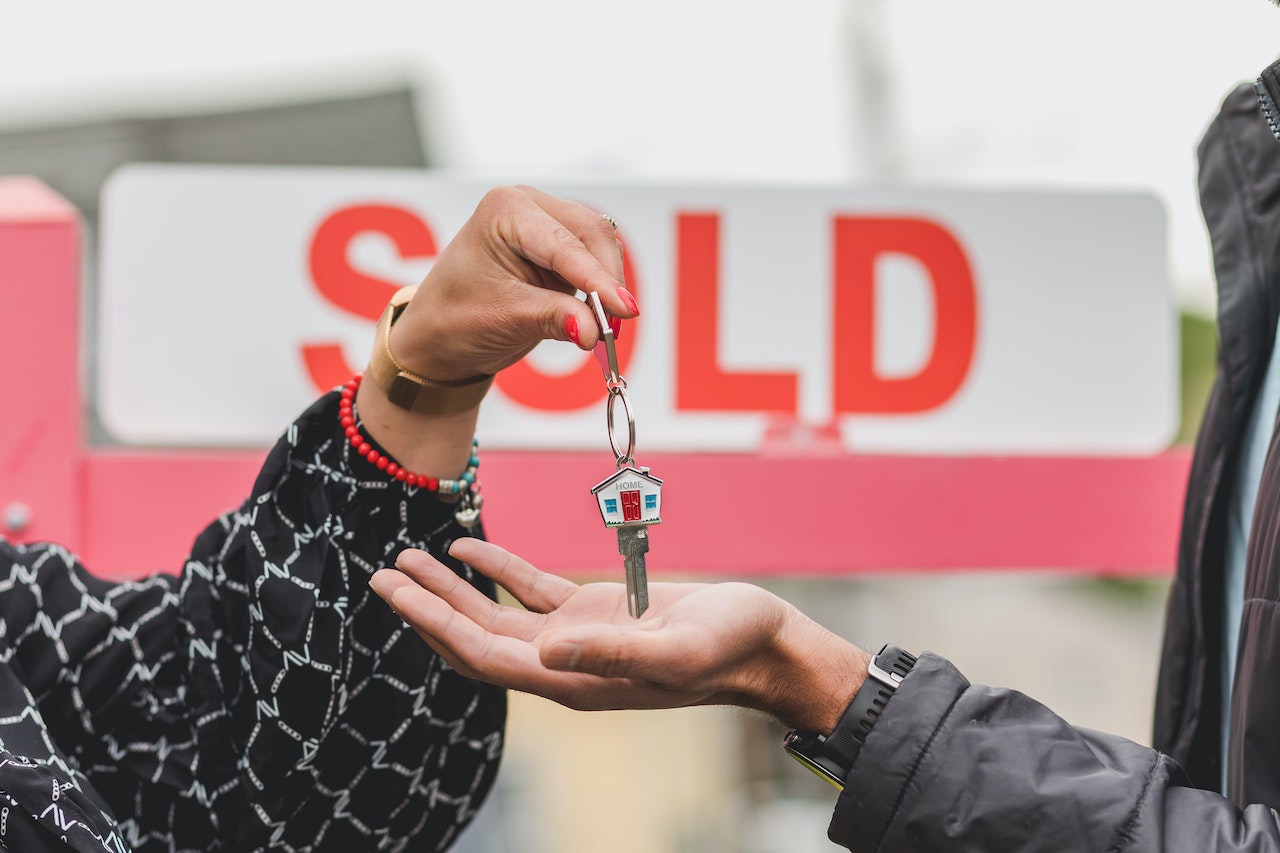 Hand holding house keys in foreground and sold sign in background.