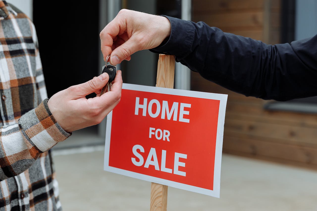 One hand giving key to another in foreground; Home For Sale sign in background. 