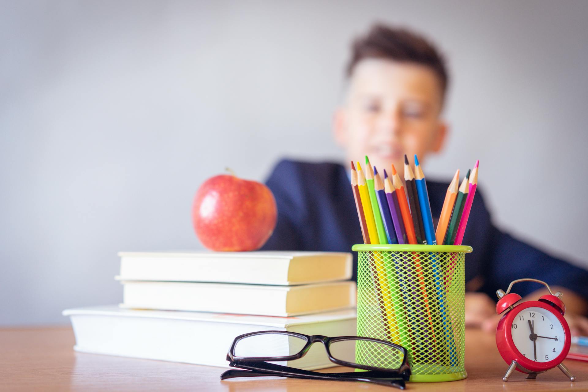 Books, pencils, apple in foreground; blurred child in background.