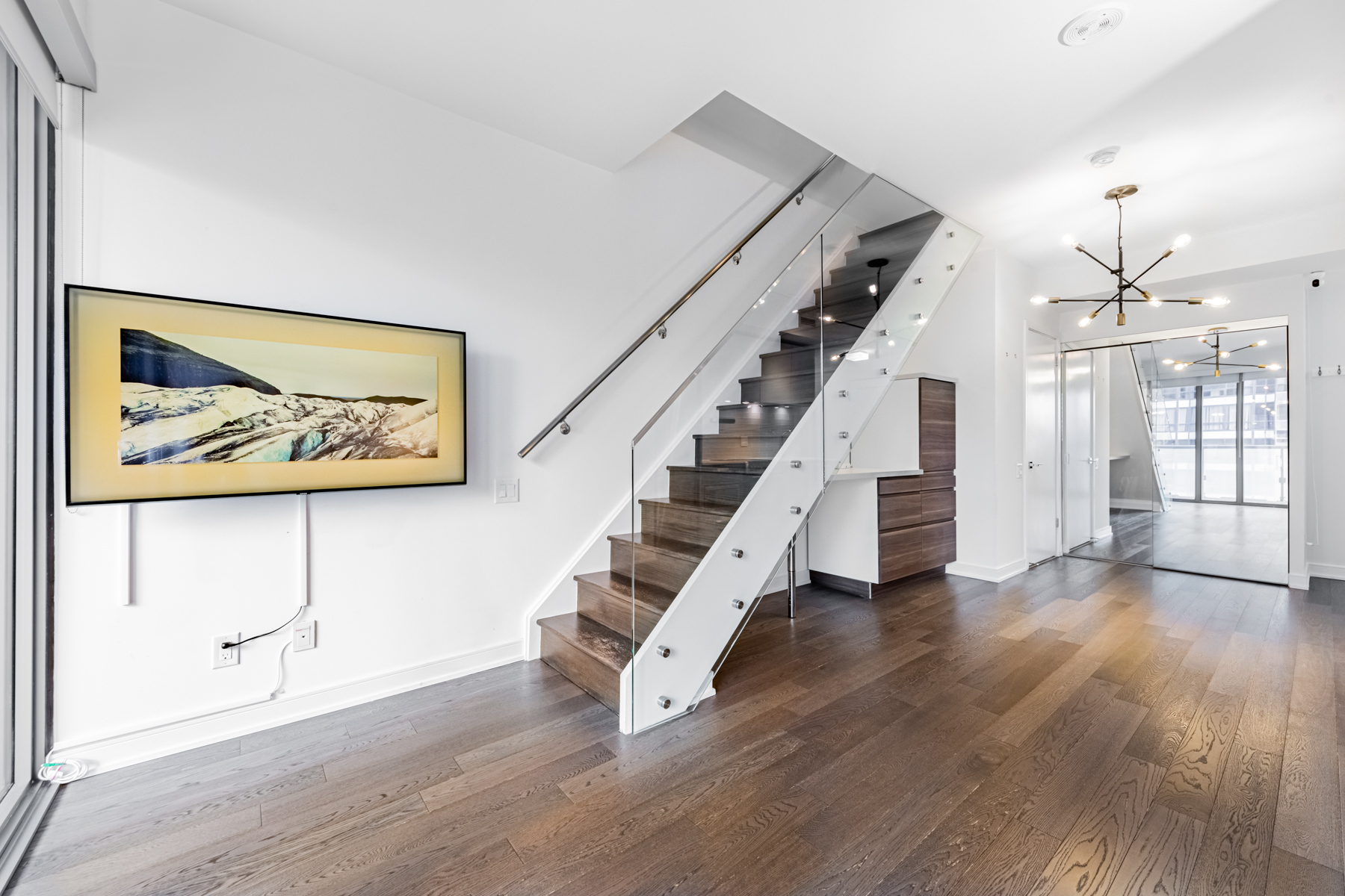 Condo with dark hardwood floors, glass banister staircase and trendy chandelier.