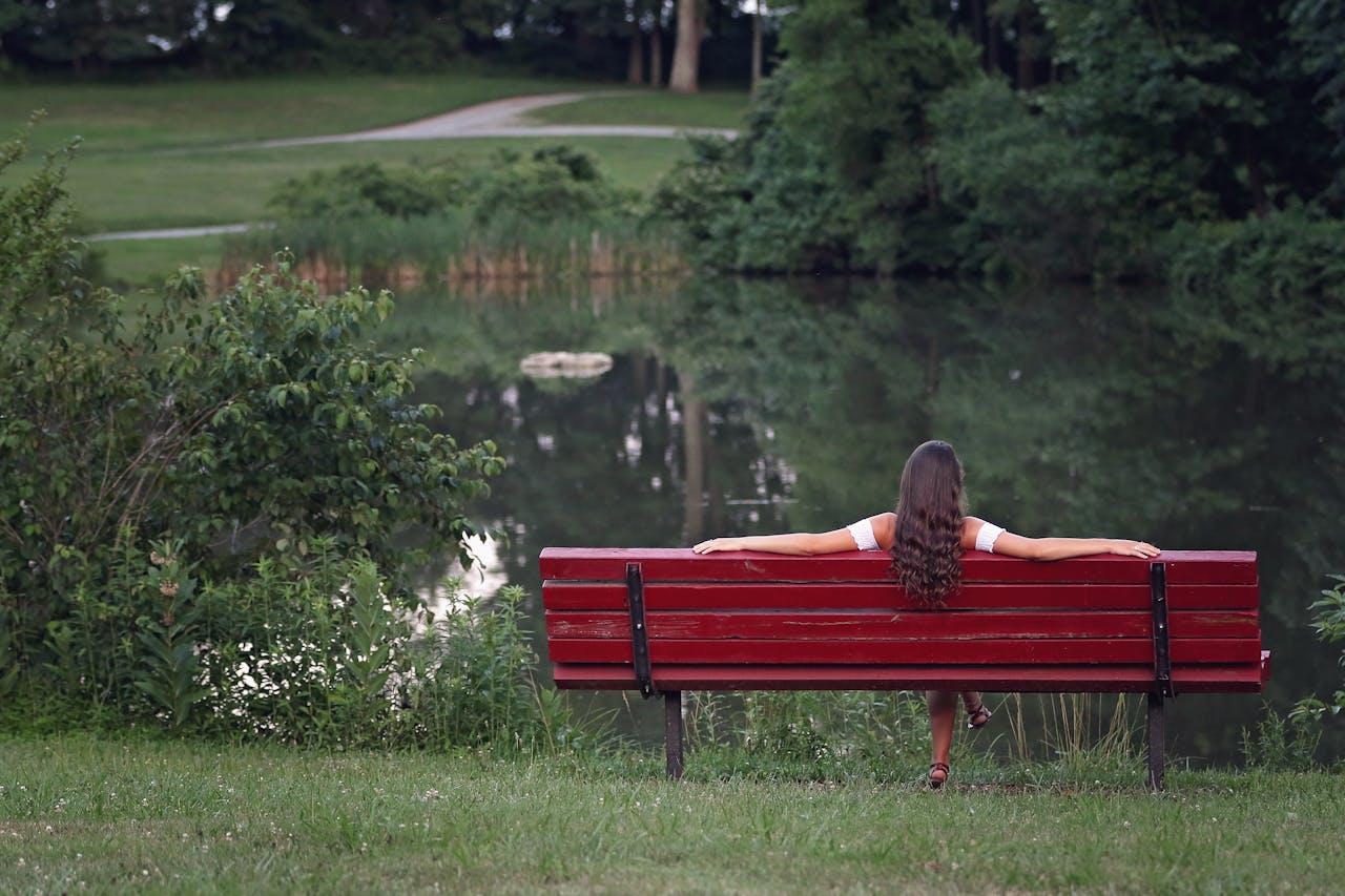 Woman sitting on red park bench in front of pond.