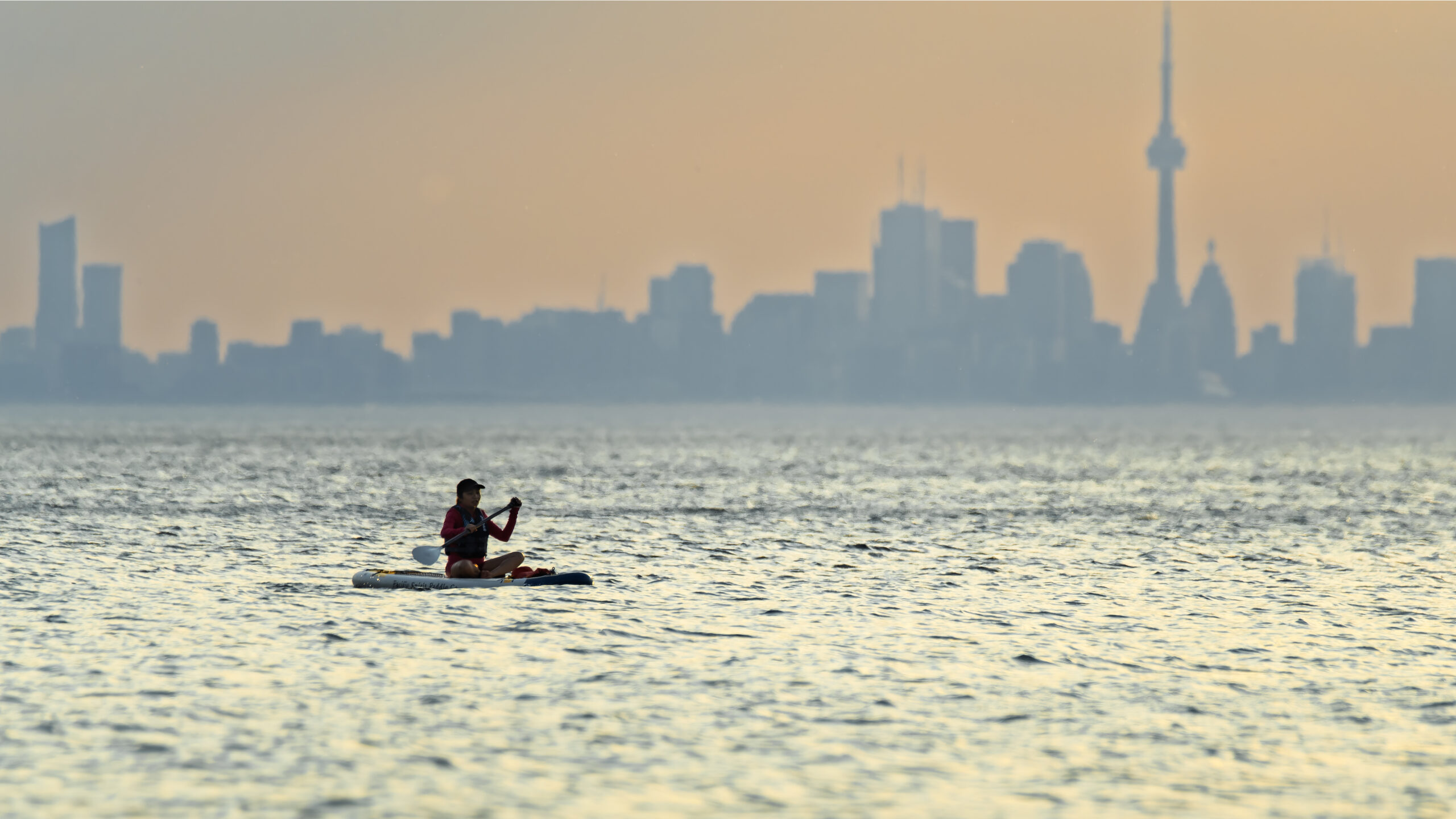 Long distance photo of woman in kayak with hazy Toronto skyline in background.