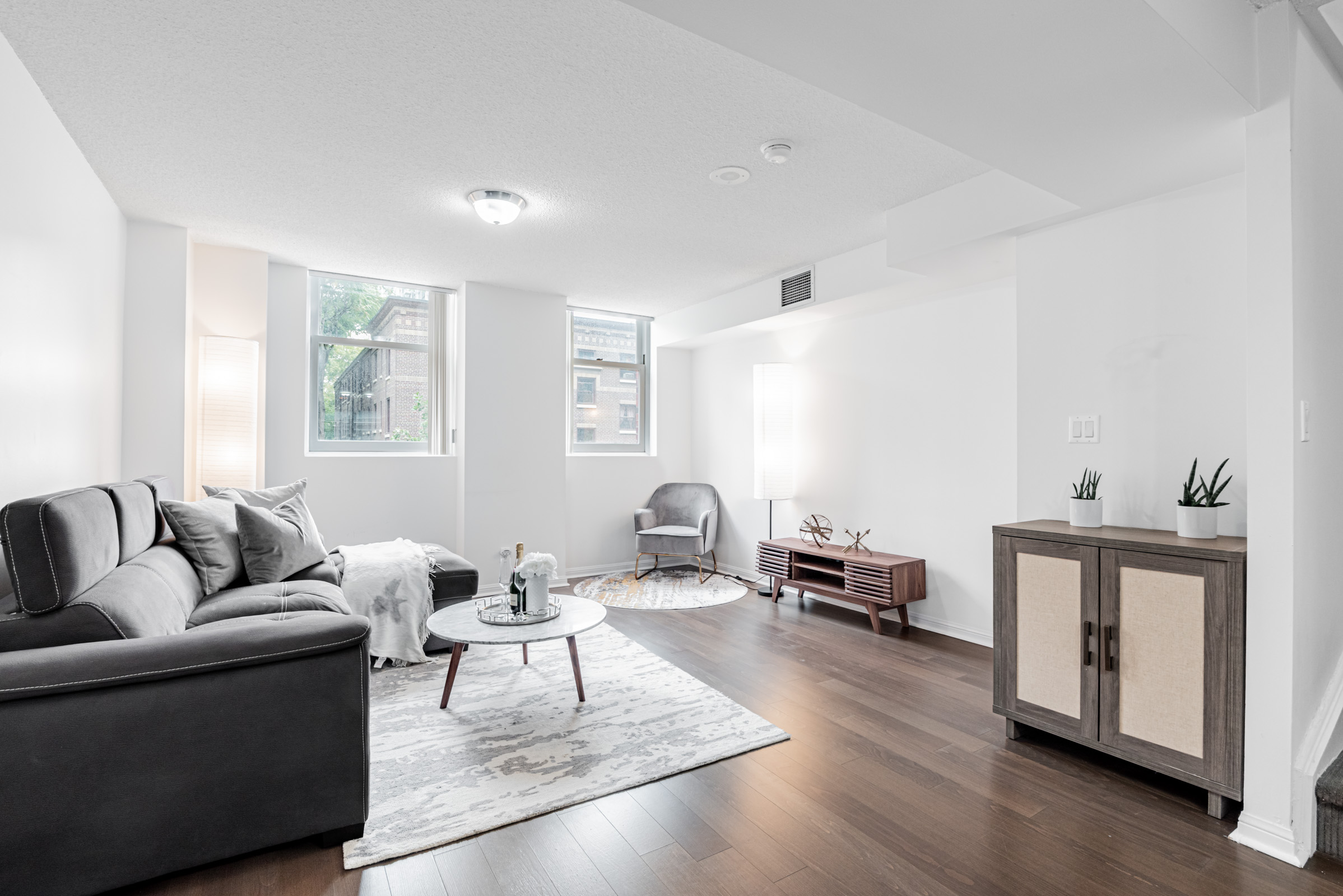 Large condo living room with dark brown laminate floors, two windows and several pieces of furniture.