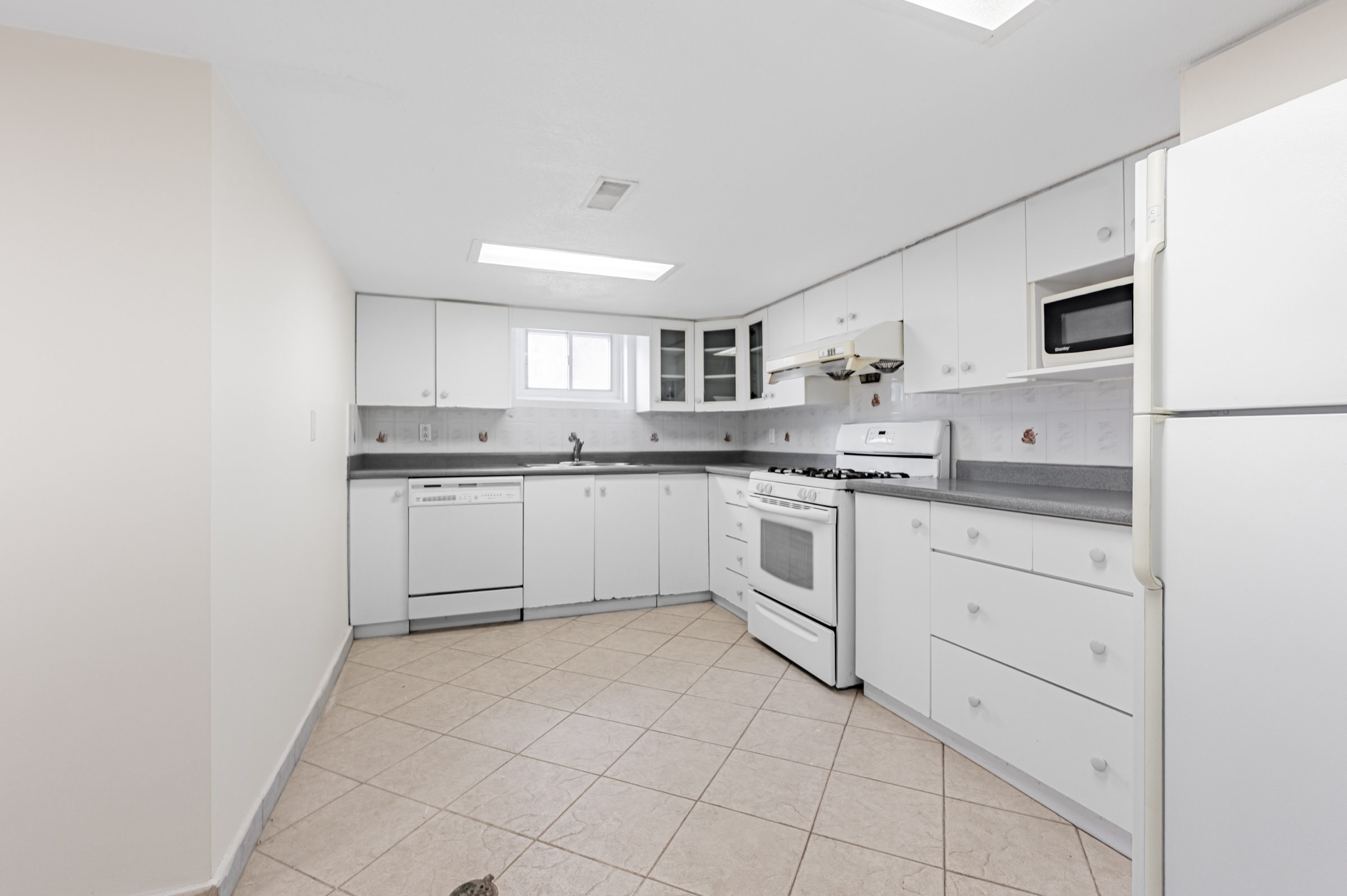 L-shaped basement kitchen with several cabinets, drawers and counters, ceramic floors, fluorescent lighting, and window.