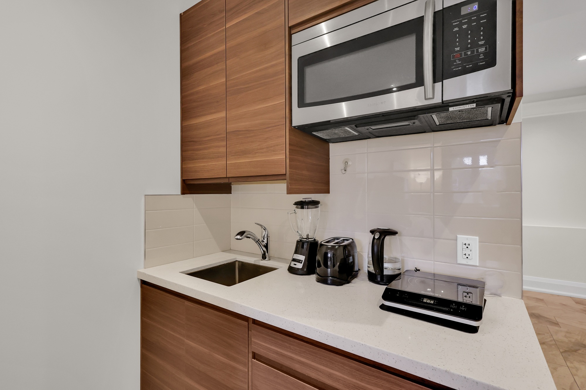 Basement kitchenette with tiled back-splash, sink, storage cabinets, microwave and exhaust vent.