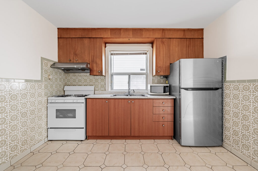 42 Carr St galley kitchen with brown cabinets and drawers, tiled floors, stainless-steel fridge, white stove, marble counter, and large window.