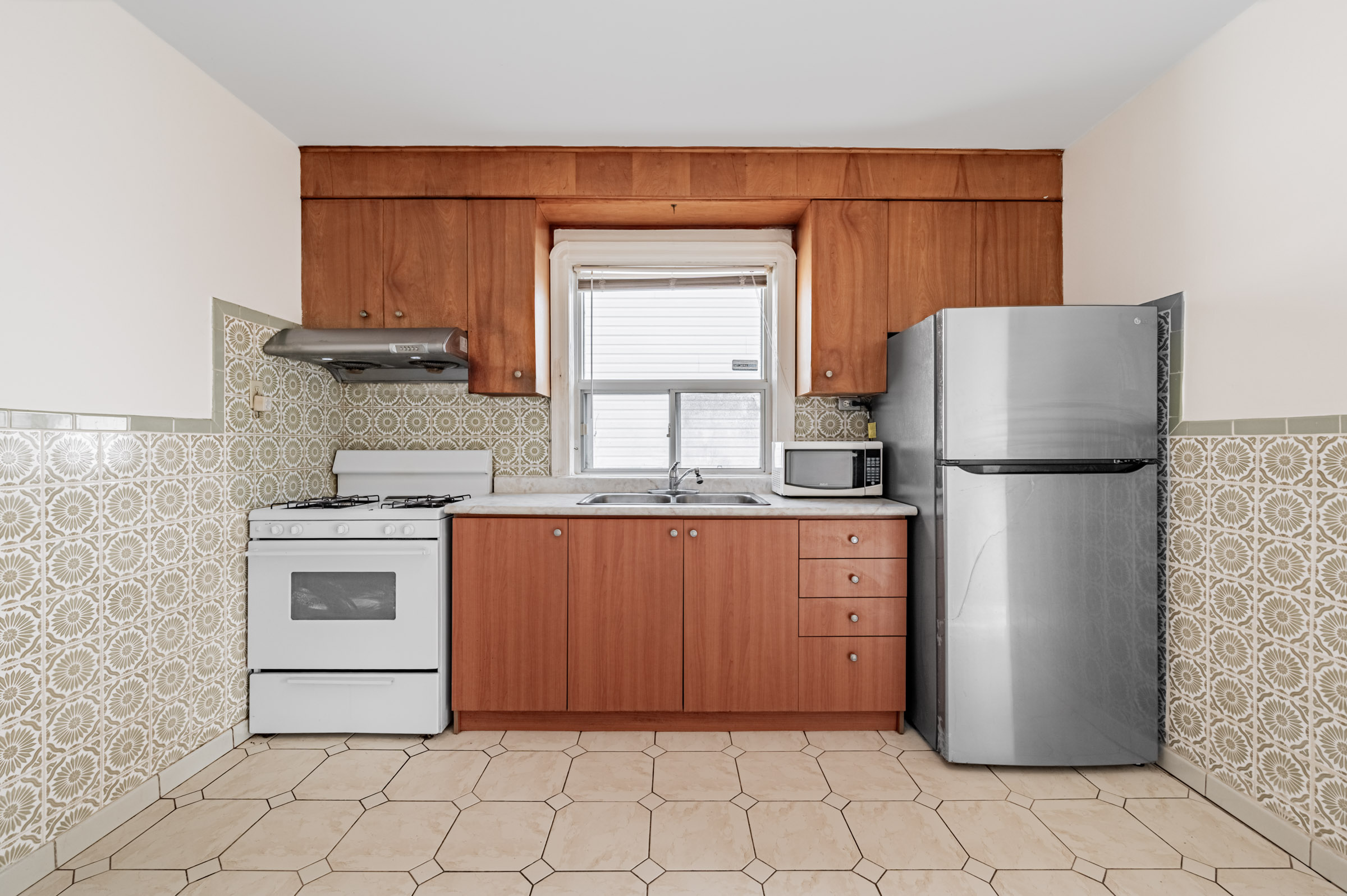42 Carr St galley kitchen with brown cabinets and drawers, tiled floors, stainless-steel fridge, white stove, marble counter, and large window.