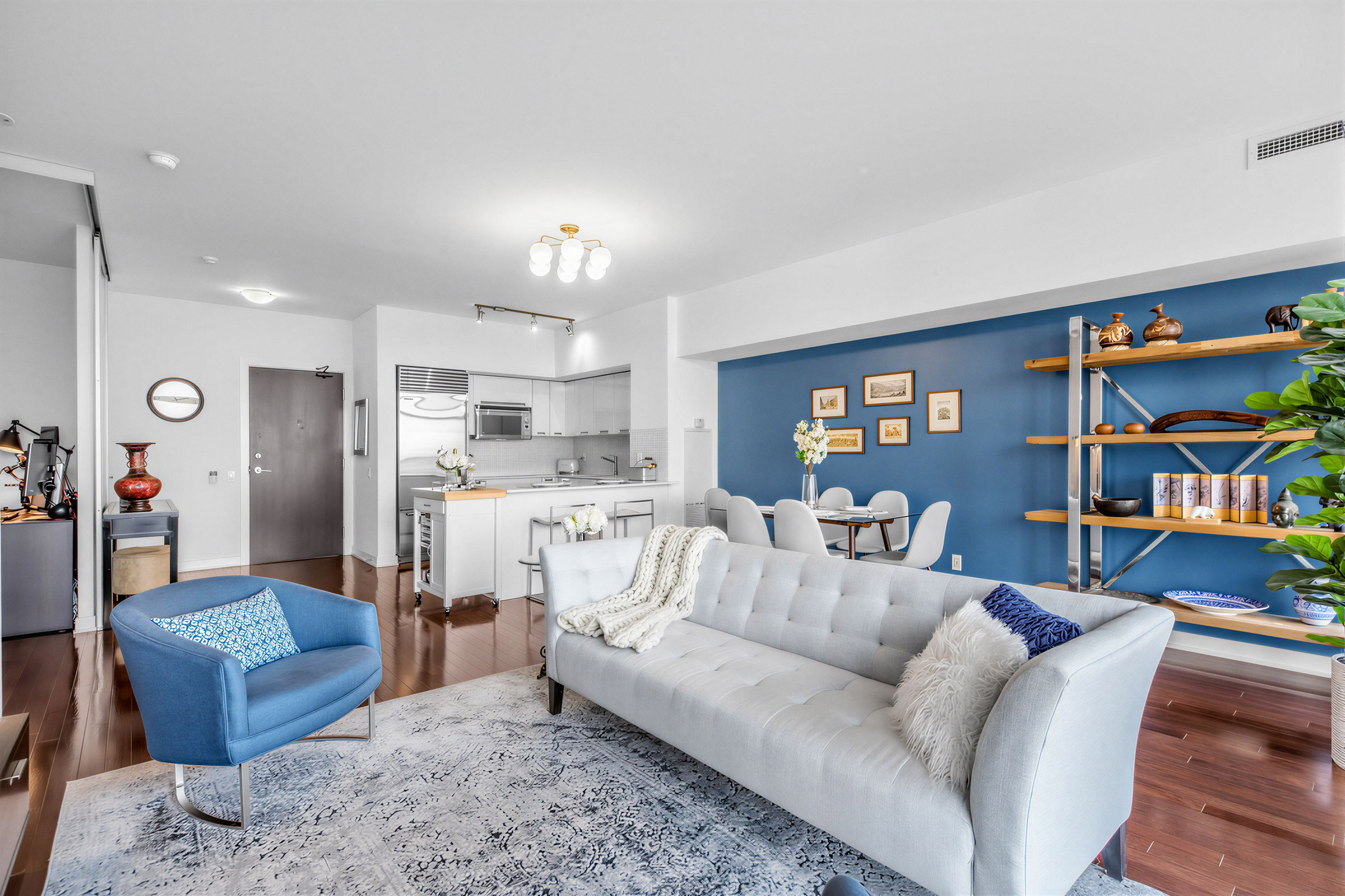 Living, dining and kitchen areas with modern light fixture, hardwood floors and blue accent wall.