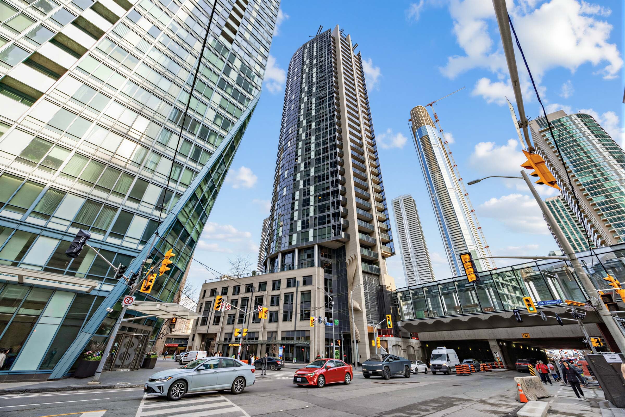 Across-the-street view of Backstage on the Esplanade condo surrounded by other buildings and with a glass and concrete pedestrian bridge over traffic. 