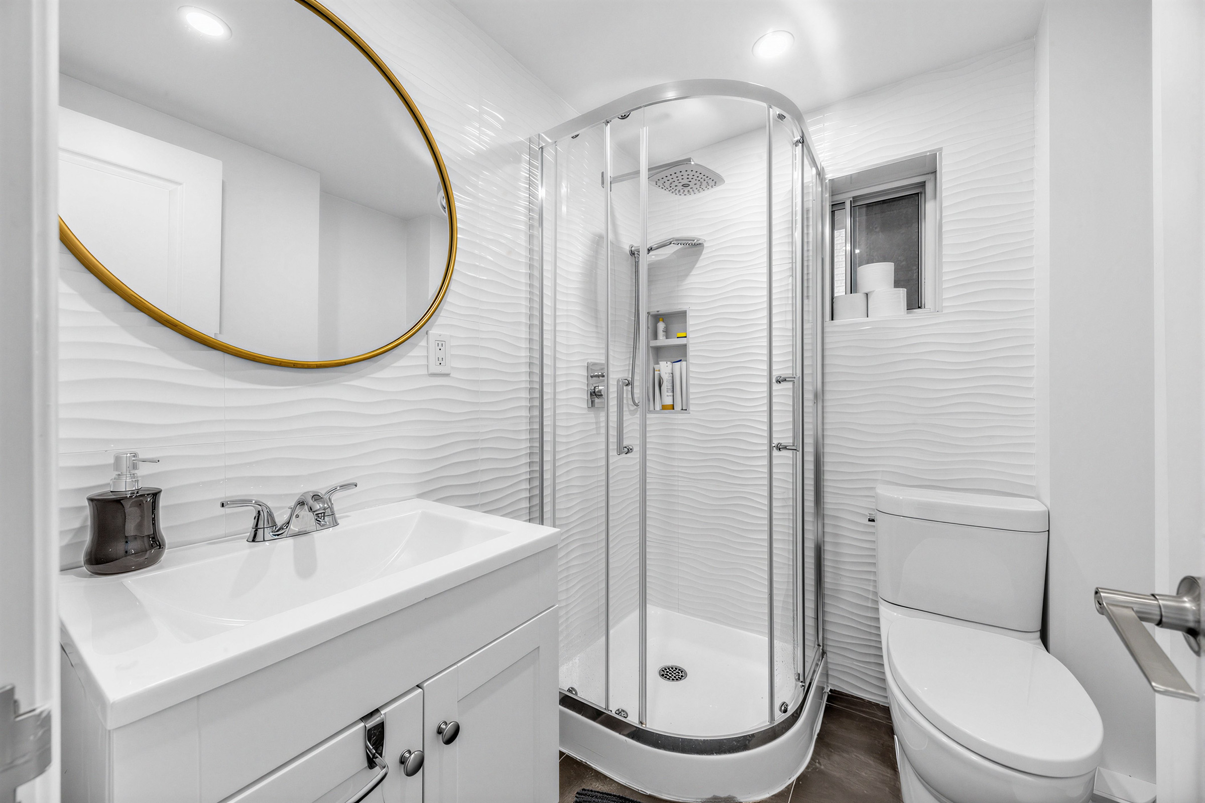 57 Shanly St main floor bath with white walls with rippling texture, gold-rimmed mirror, sink with chrome faucet, and walk-in shower with curved design and rainfall showerhead.