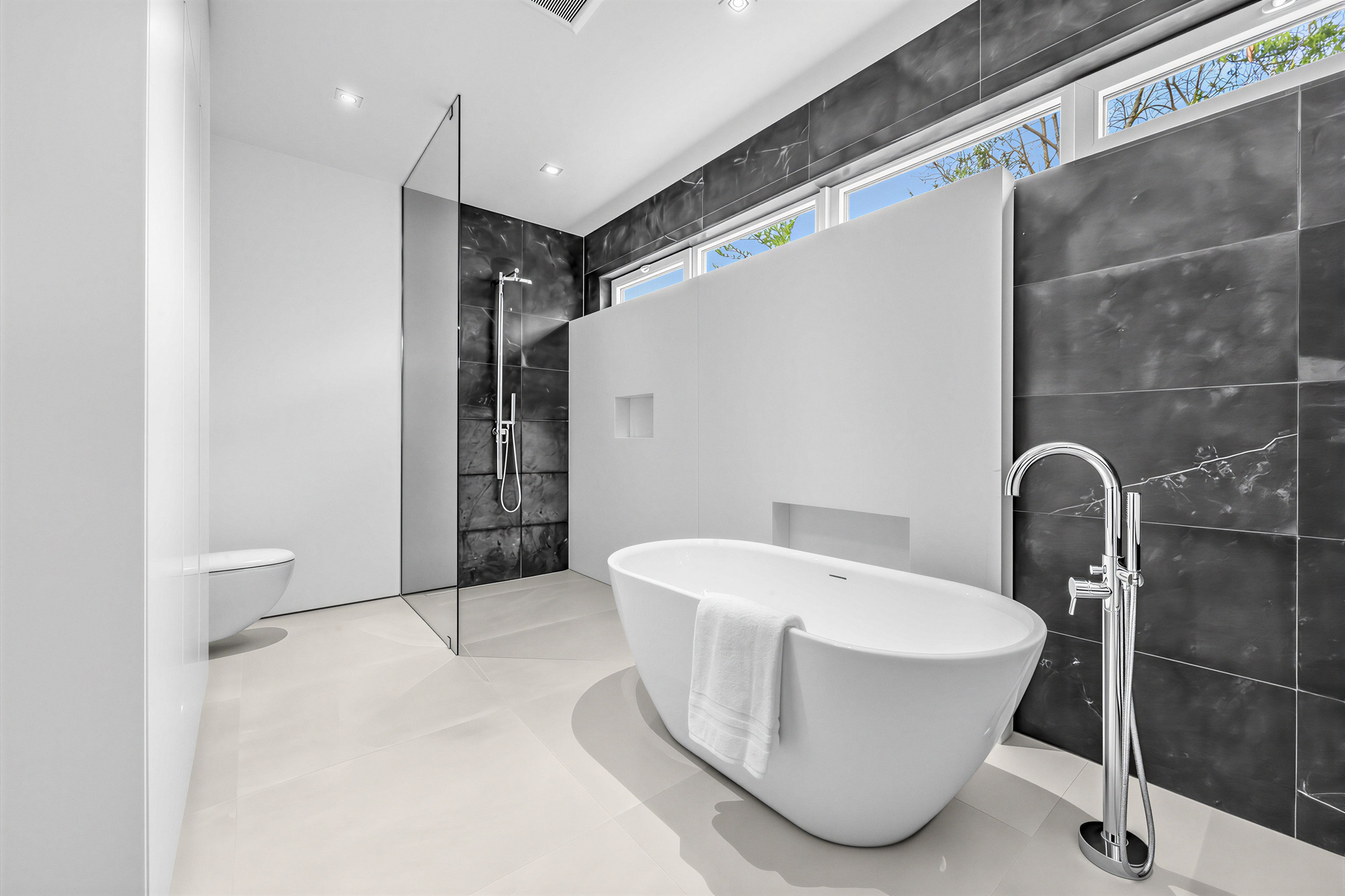 Bathroom walk-in shower with a glass partition, rainfall showerhead, and light gray bump out wall in the black tiled expanse.