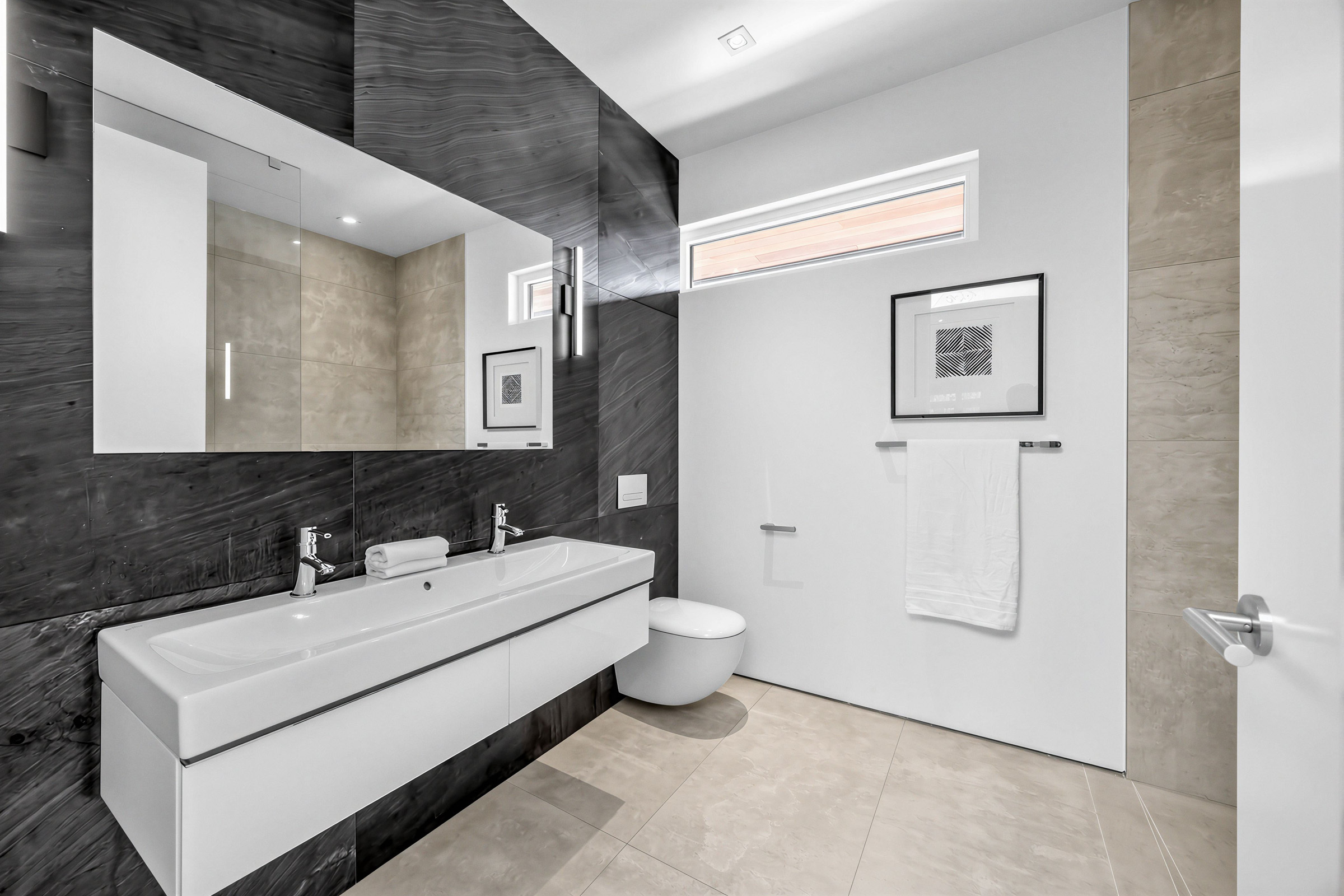 4-piece bath with black tiled wall, floating sink with dual faucets, thin wall-mounted mirror, long window and beige tiles.