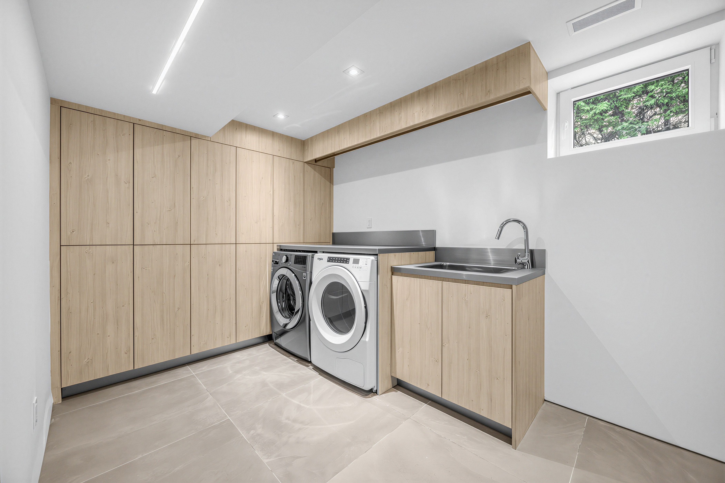 Basement laundry room with washer and dryer, stainless-steel sink, ceiling lights, and floor-to-ceiling storage cabinets.