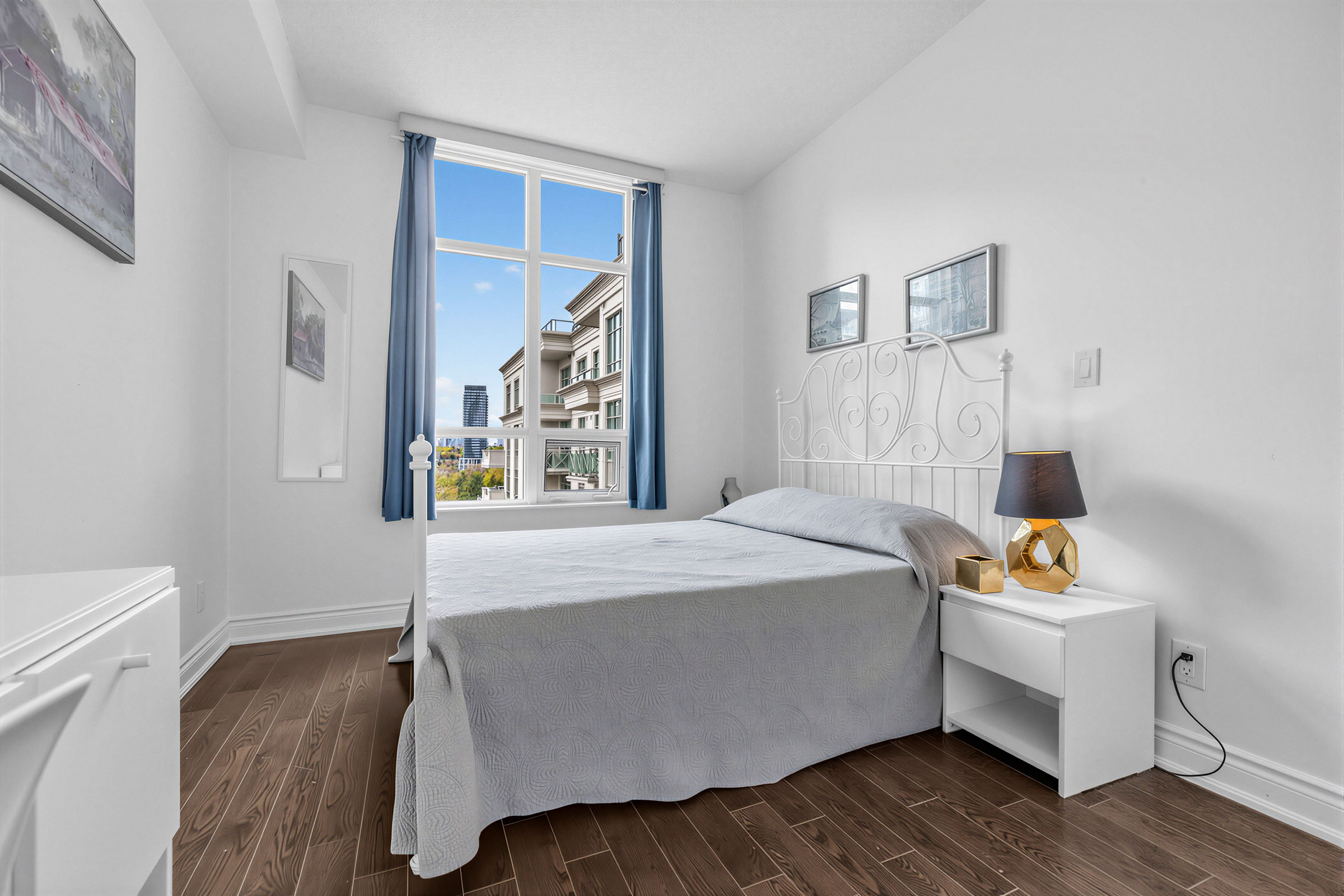 Condo bedroom with large bed, dark hardwood floors and tall window with view of building.