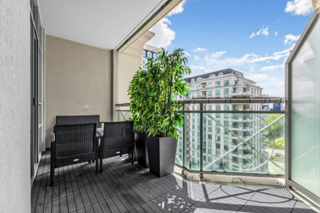 20 Bloorview Place Unit 1003 balcony with black chairs, black floor, potted plants, glass panels, glazed divider, and view of building and skies.
