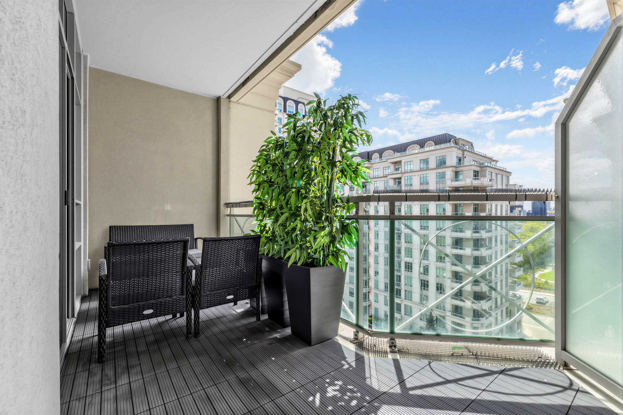 20 Bloorview Place Unit 1003 balcony with black chairs, black floor, potted plants, glass panels, glazed divider, and view of building and skies.