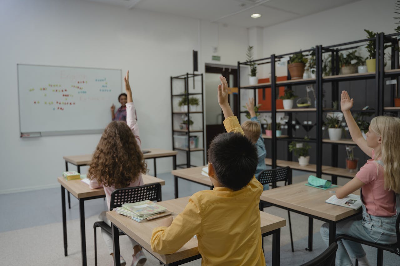 Kids in classroom raising hands as teacher stands at front of class.