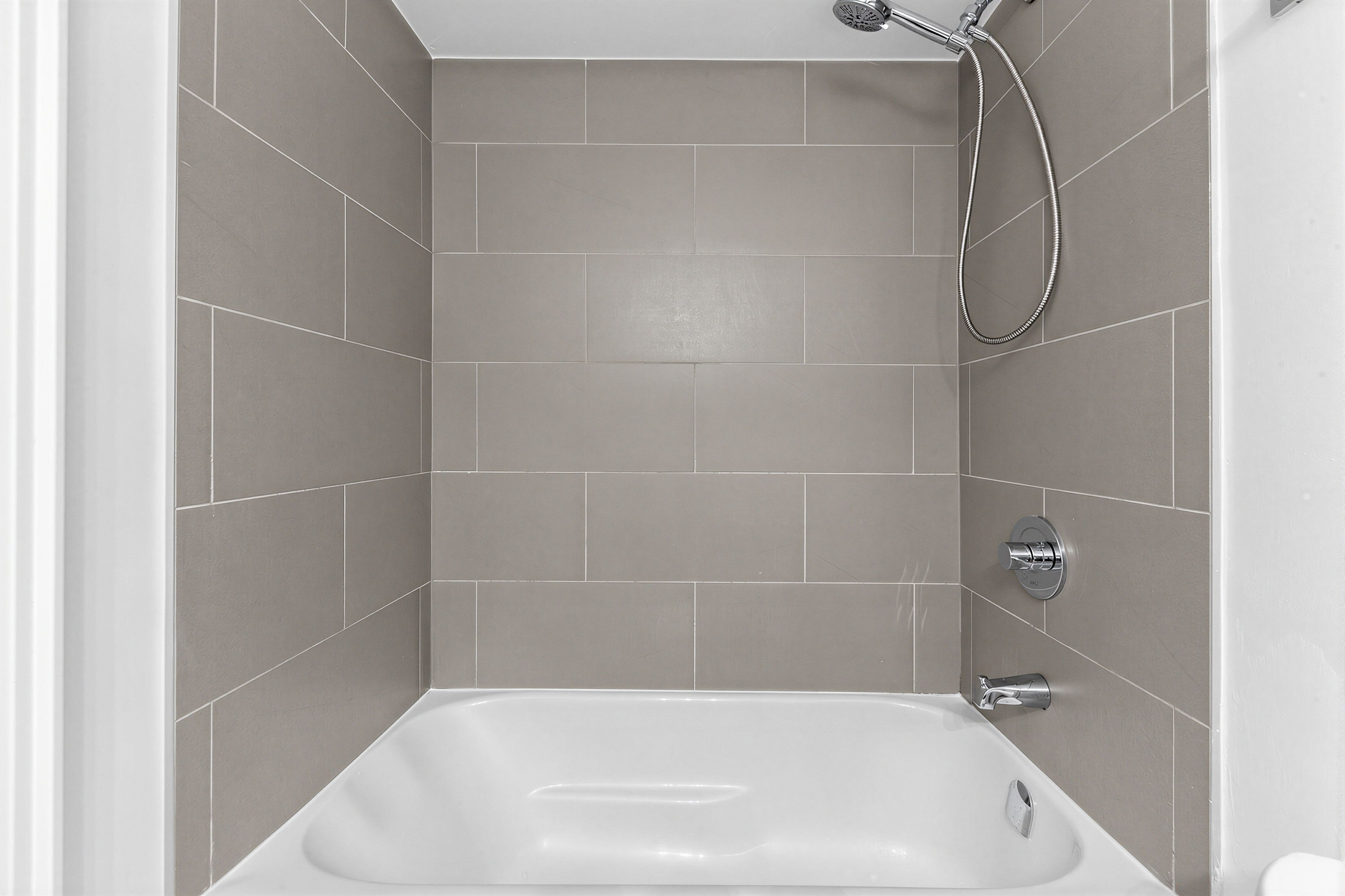Condo bath with white tub, walls with large ceramic tiles, and detachable showerhead.