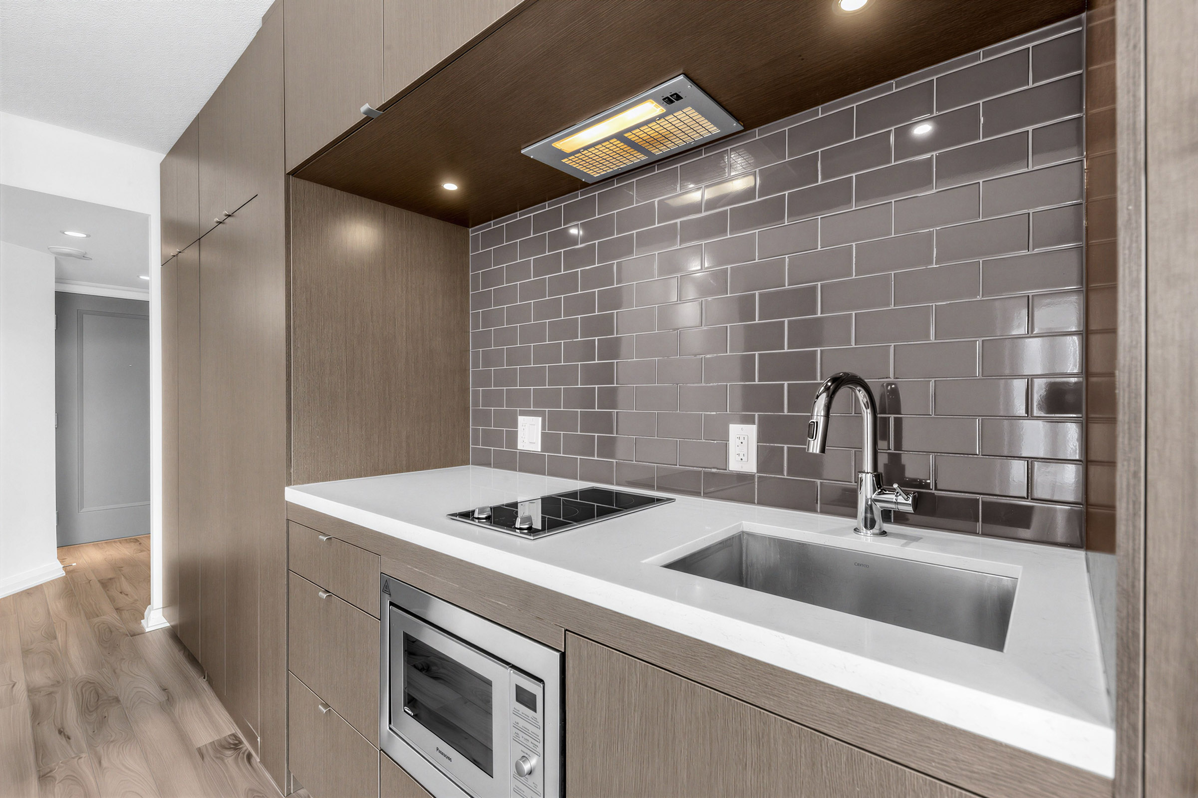 Condo kitchen with dark grayish-brown subway tile backsplash, white quarts counters, LED downlights, and range hood panel lighting.